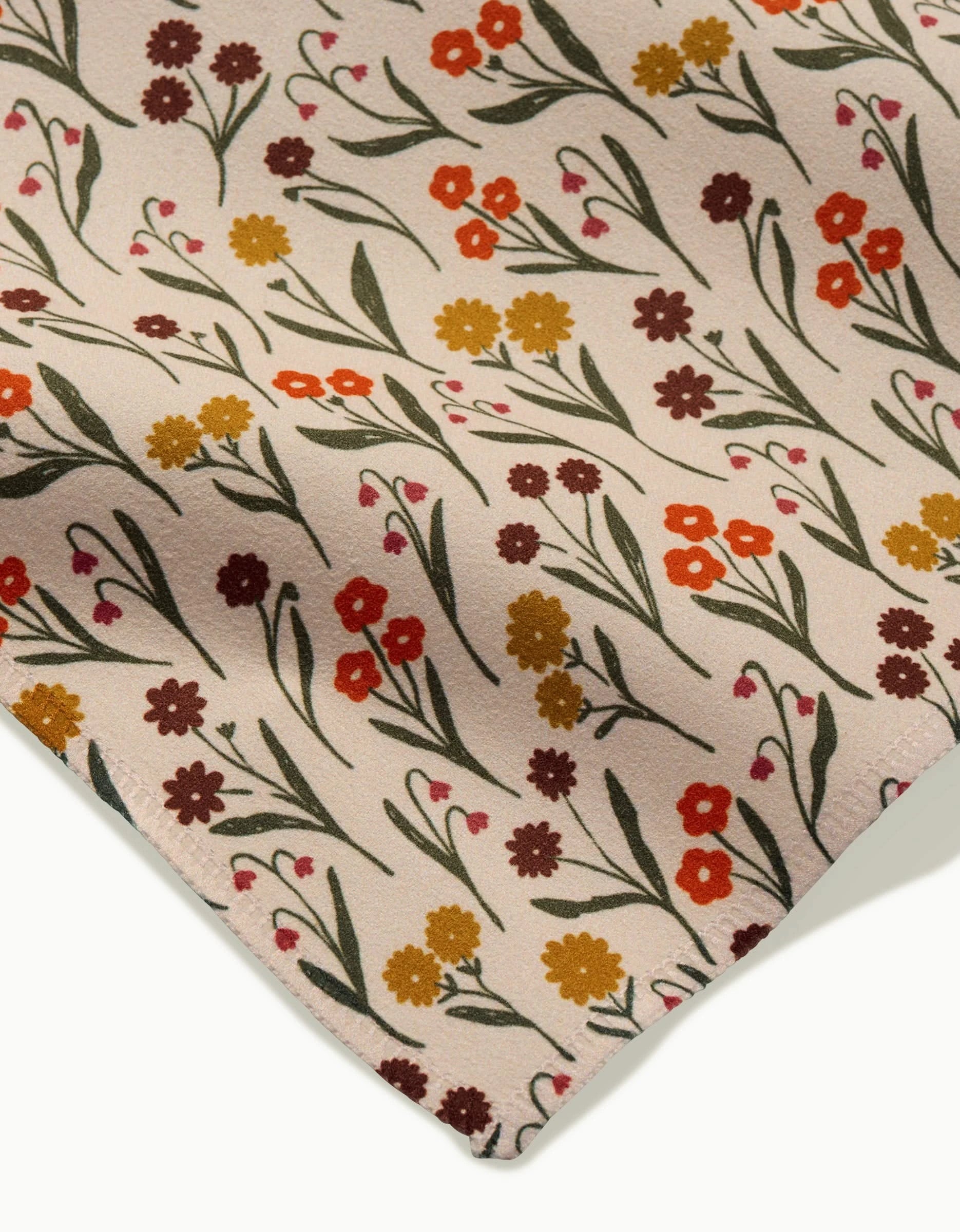 A close-up of Kayla Ann’s Fall Blossom fabric, showing a floral pattern with small red, yellow, and brown flowers with green stems and leaves on light beige Geosuede. The fabrics edge appears in the lower corner.