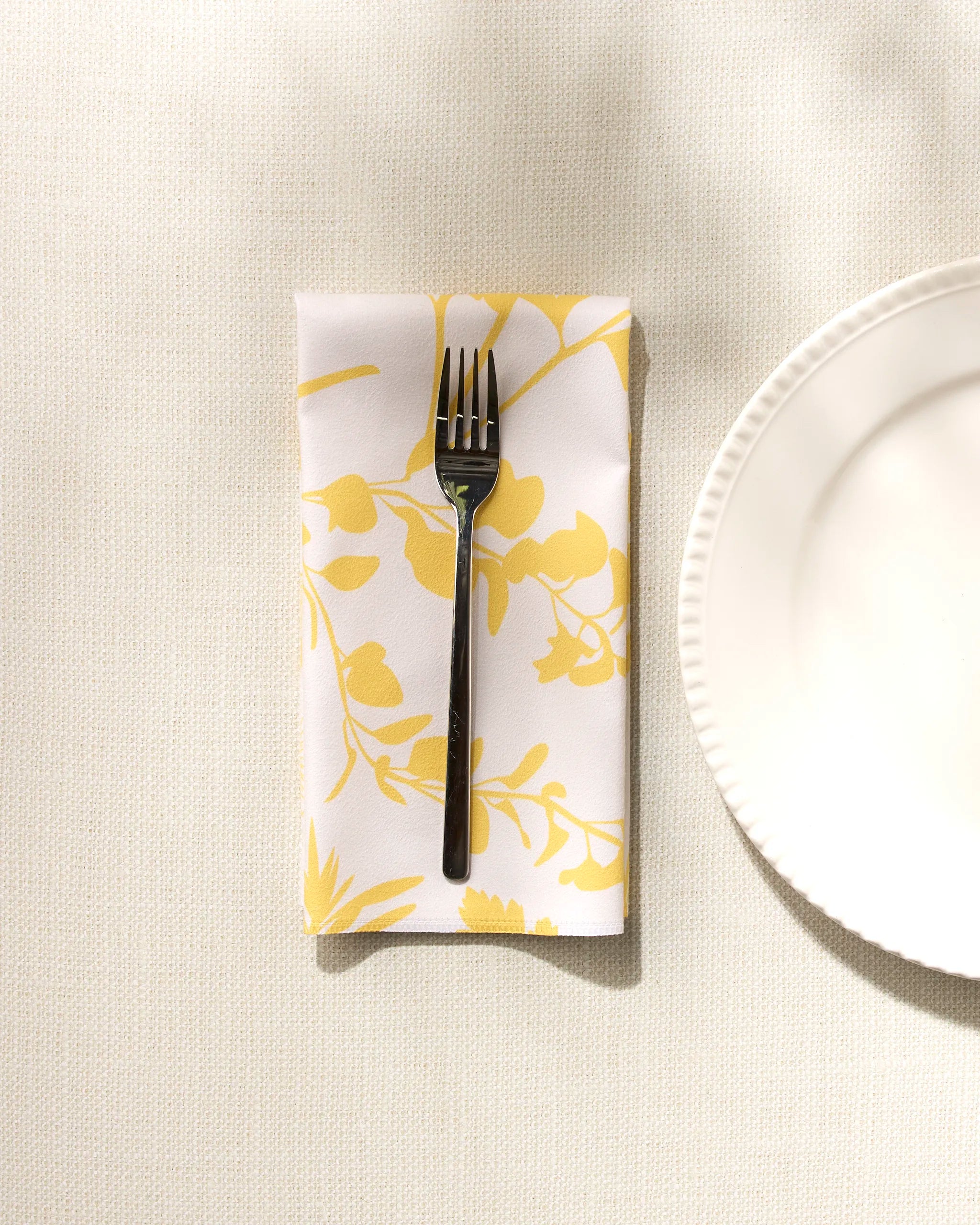 A silver fork rests on GEOMETRY's "Garden of Gold" yellow and white floral washable napkin, folded beside a white plate on a textured, light-colored tablecloth.