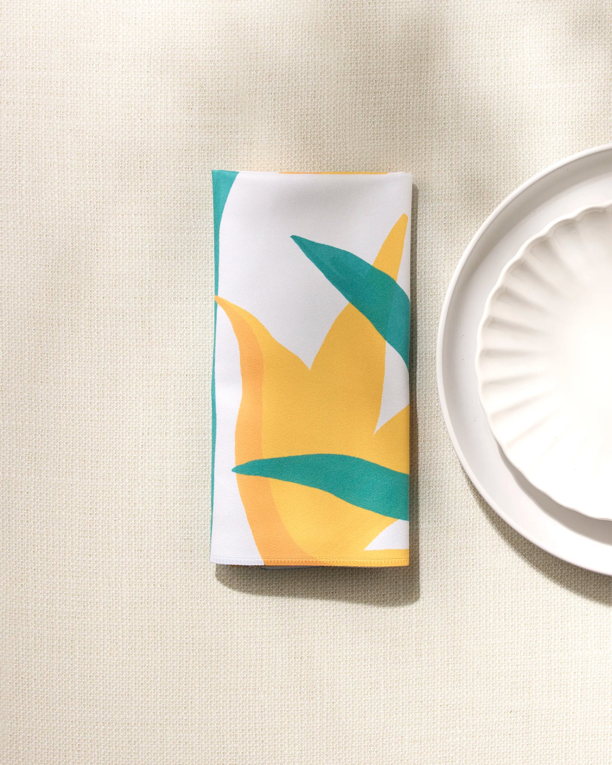 The GEOMETRY "Growth" napkin, featuring a yellow and green abstract leaf design on white, is neatly folded beside a white scallop-edged plate on a textured cream surface.