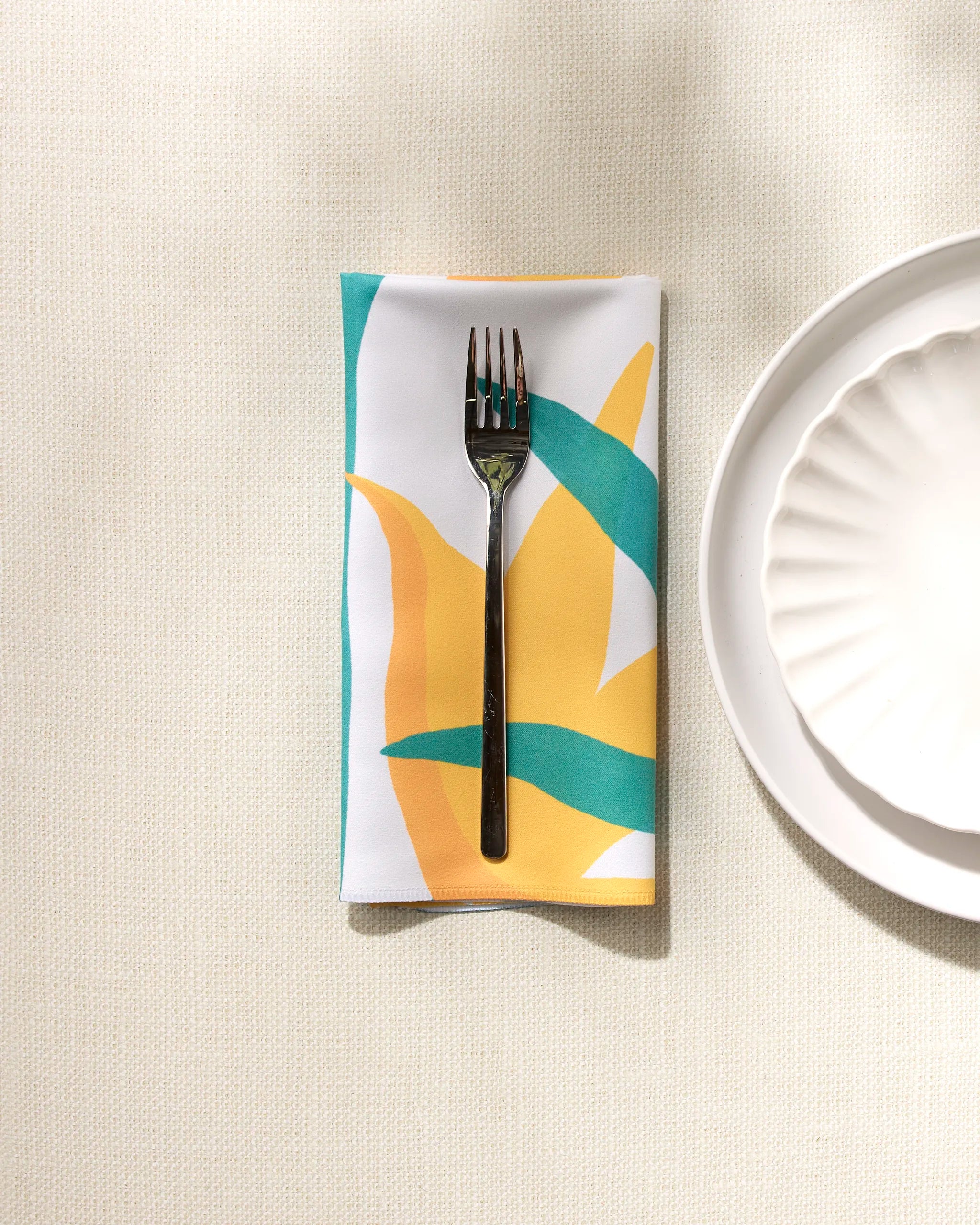 A fork rests on a folded GEOMETRY "Growth" recycled napkin with green, yellow, and orange shapes beside a white scalloped plate atop a textured light tablecloth.