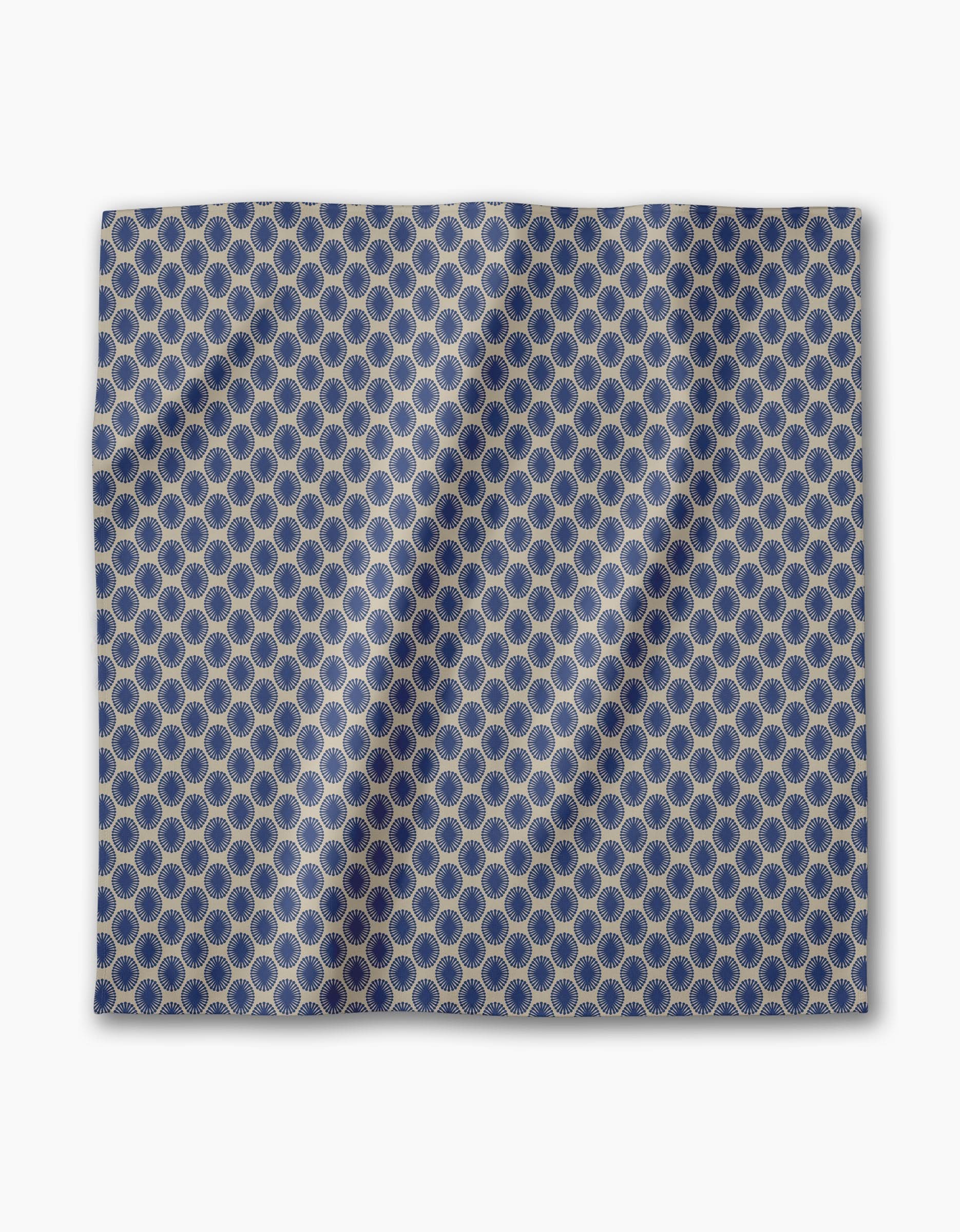 The Lagoon dinner napkin by Geometry is a blue fabric square with interlocking gold hexagons, shown flat with subtle folds and shadows. This absorbent cloth napkin is part of an elegant dining set.