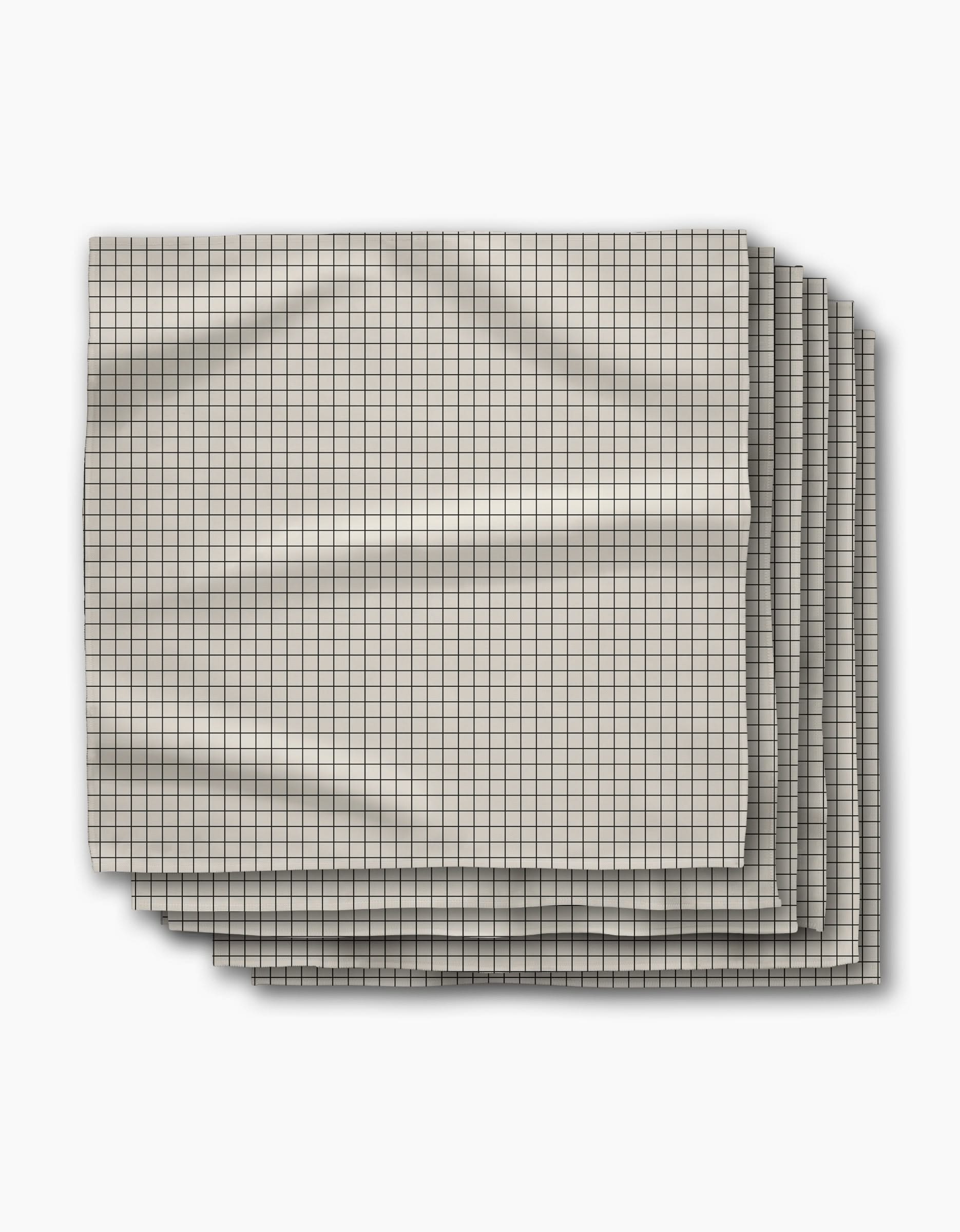 Four folded Metro Grid dinner napkins by Geometry, featuring a small black grid pattern on light beige Geosuede fabric, are stacked slightly offset on a white surface with visible creases.