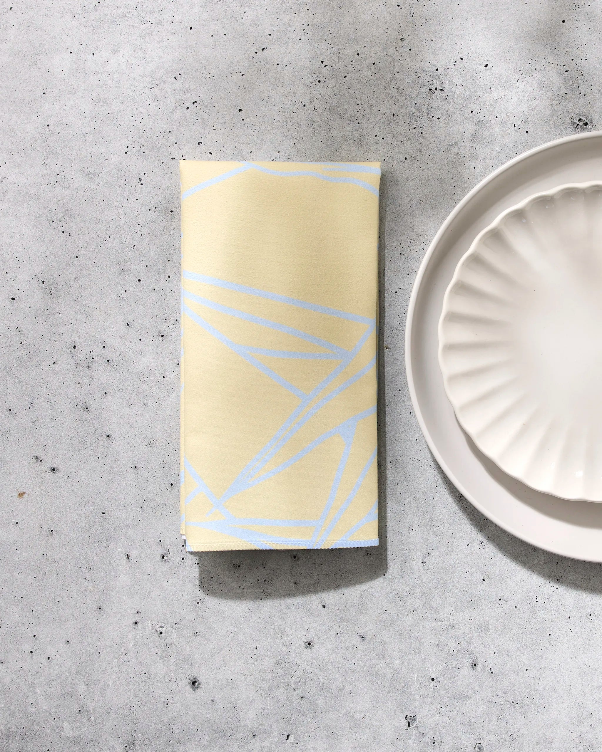 The GEOMETRY Paradise Blooms folded yellow washable napkin with light blue geometric lines rests on a gray speckled surface beside two stacked white scalloped-edge plates.