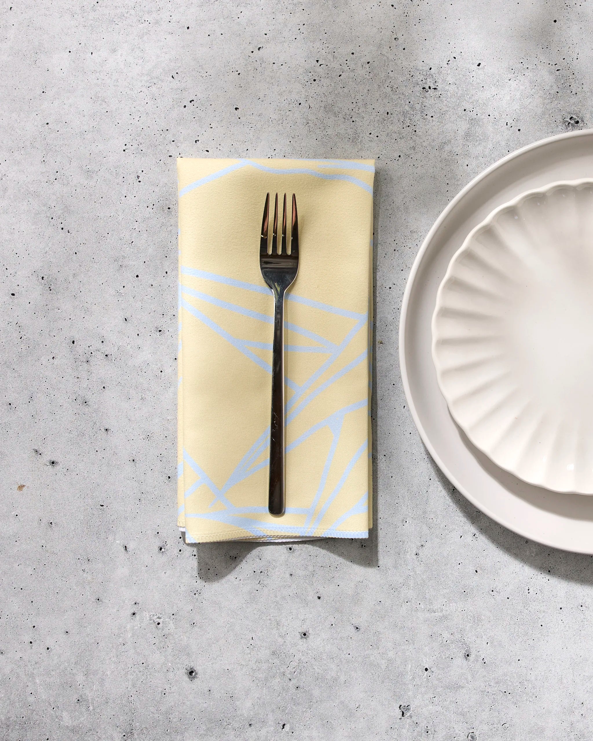 A GEOMETRY Paradise Blooms fork sits on a folded, patterned yellow dinner napkin beside two stacked scalloped white plates on a light gray textured surface.