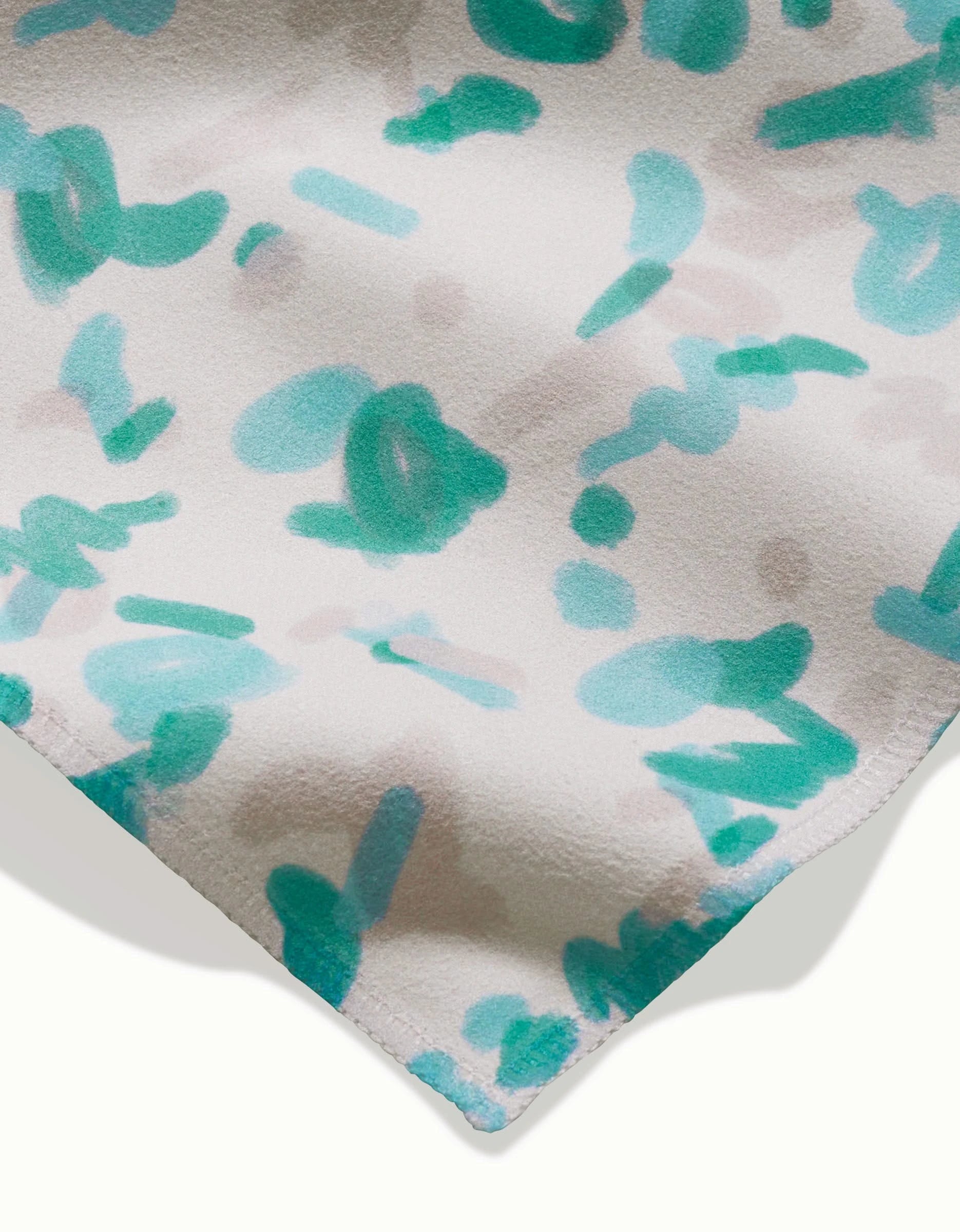 A close-up of GEOMETRY’s “Playful Blues” napkin shows eco-friendly Geosuede fabric with teal brushstrokes and pale gray spots on white, plus a stitched edge—ideal for dinner napkins.