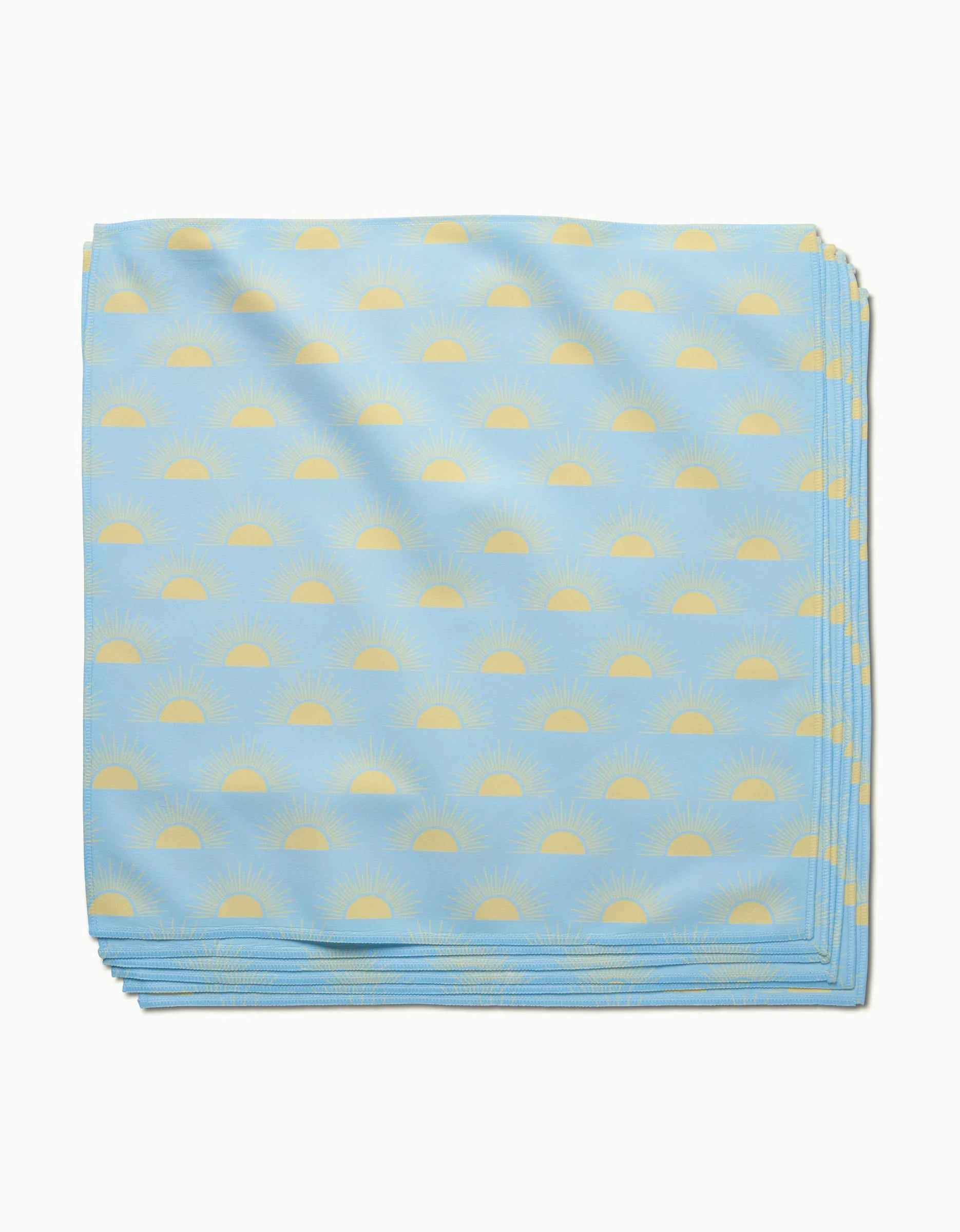 The GEOMETRY Soft Summer Sun dinner napkin, folded from light blue Geosuede fabric with a minimalist pattern of yellow sunrises and radiating rays, is neatly stacked against a white background.