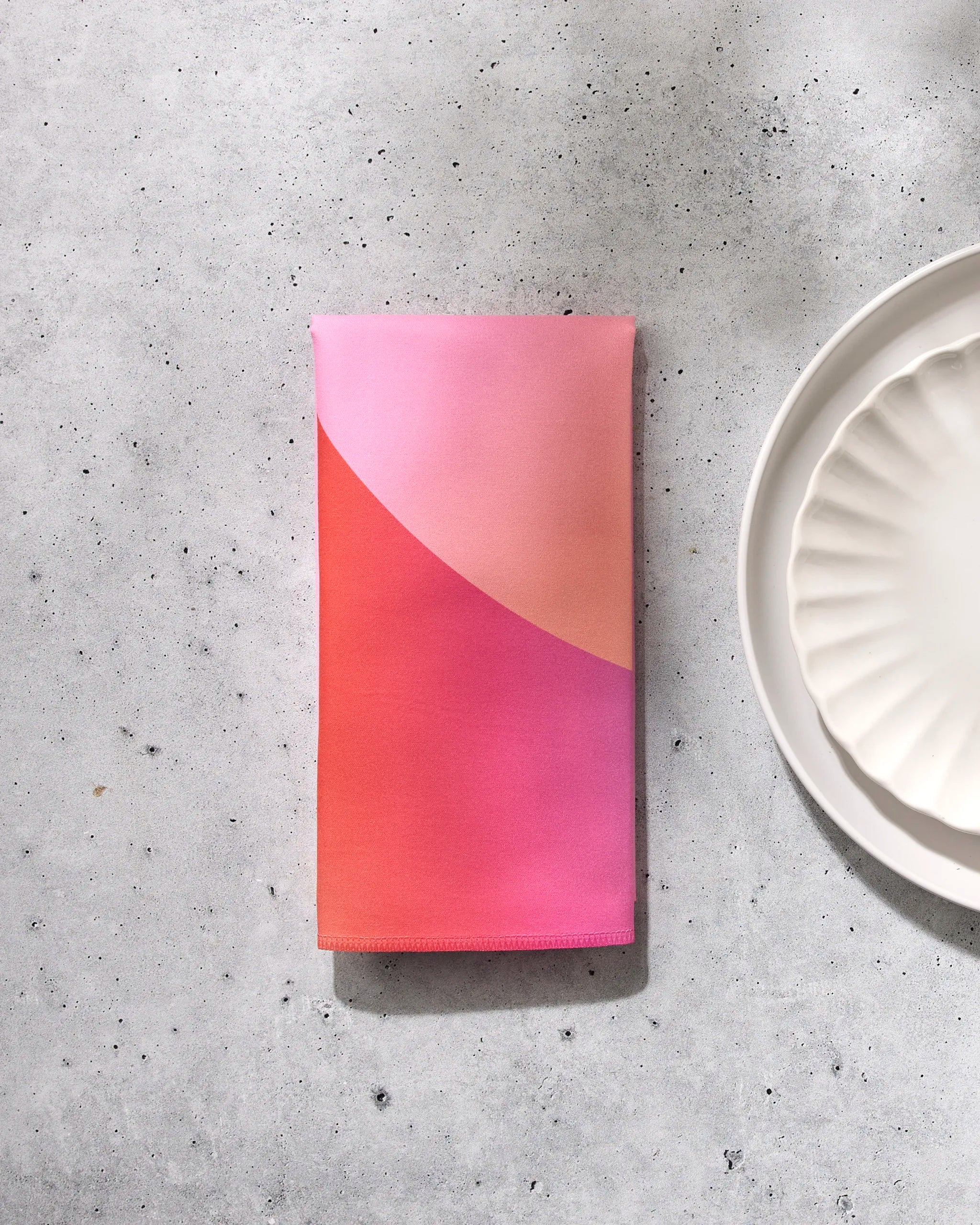 The Sunrise eco-friendly napkin by GEOMETRY, featuring a pink and orange gradient, is folded on a light speckled surface beside a scalloped white plate, with soft shadows completing the scene.
