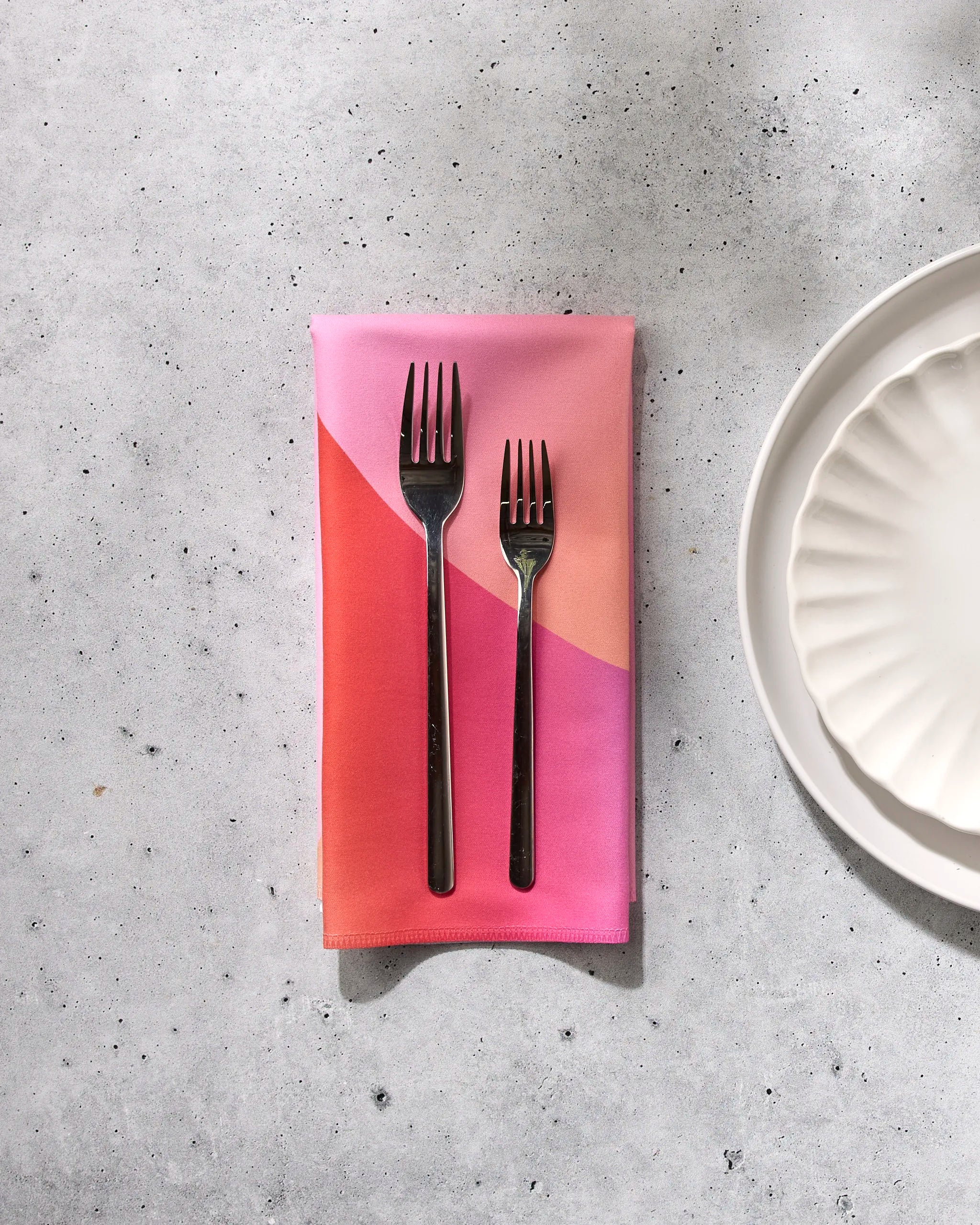 Two GEOMETRY Sunrise forks rest side by side on a folded eco-friendly napkin in pink and orange, next to a white scalloped plate atop a light gray speckled surface.