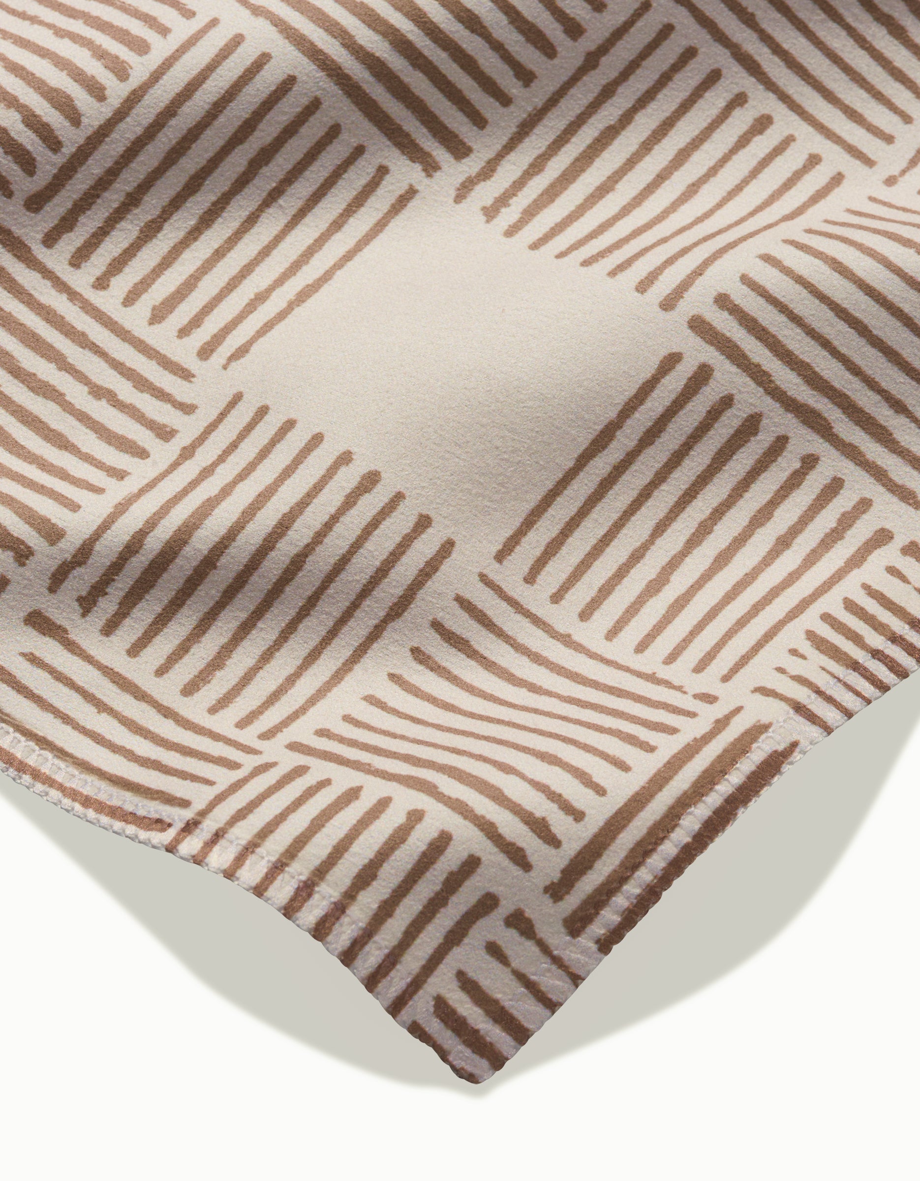Studio Jeannots Thatcher features beige geosuede with a geometric pattern of brown parallel lines forming diamond shapes—shown in close-up with soft shadows and a curved edge; perfect for an elegant dinner napkin set.