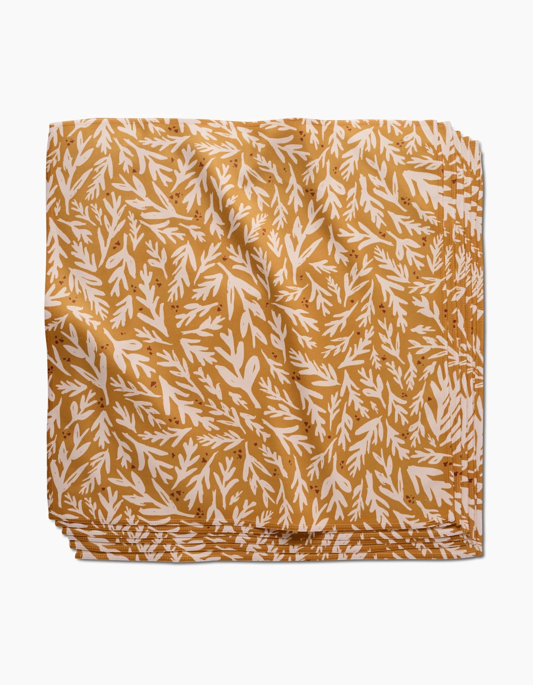 The Trail Dusting Gold napkin by Twig + Spark features an absorbent, golden yellow cloth with a white leafy branch pattern, neatly folded and stacked on a plain white surface.