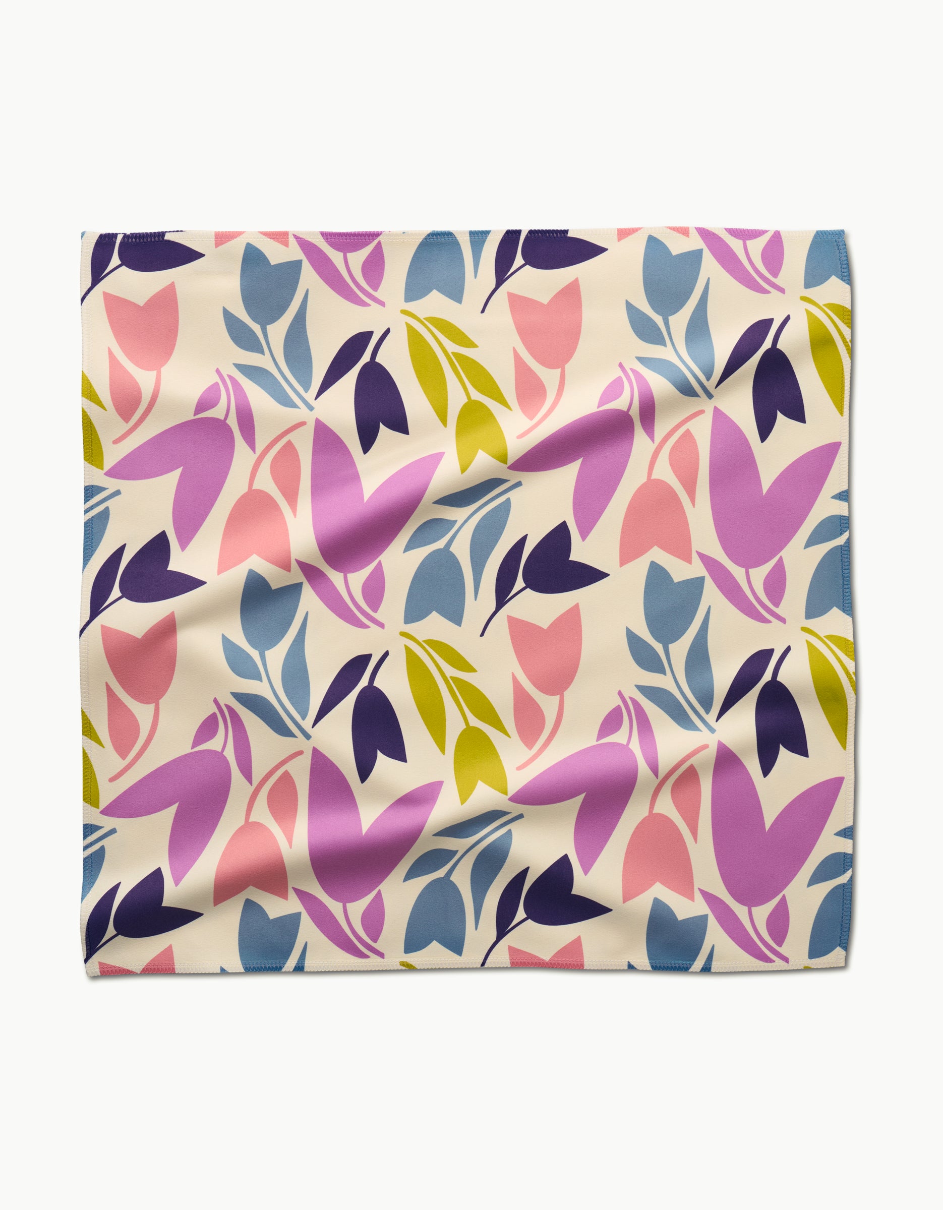 The Tulip Tumble dinner napkin by Lizzie Clark features a colorful abstract floral pattern with pink, purple, blue, and yellow leaves and petals on cream Geosuede fabric. The rectangular napkin is shown laid flat with slight wrinkles.
