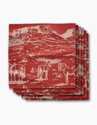 Geometry’s “Western Scene” napkin is a red, recycled dinner napkin featuring a white line drawing of mesas, horses, and desert plants. The eco-friendly napkins are stacked neatly with the design prominently displayed.