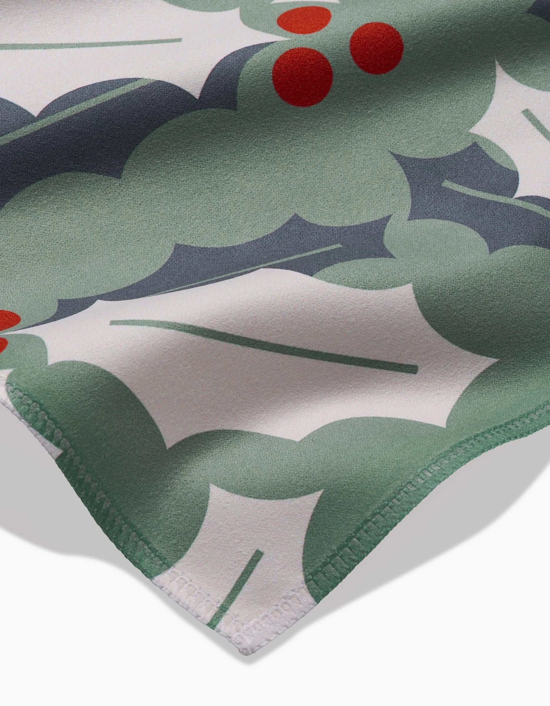 A close-up of the Lizzie Clark Winter Wonderful reusable dinner napkin shows a festive holly pattern with green leaves, white accents, red berries, smooth fabric texture, and neatly stitched edges.