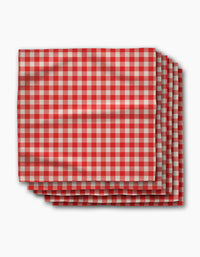 Four folded Ripe Tomato Gingham napkins by Geometry, featuring a red and white checkered pattern, are stacked neatly on a white background. Made from Geosuede fabric, this set blends softness with sustainable style.