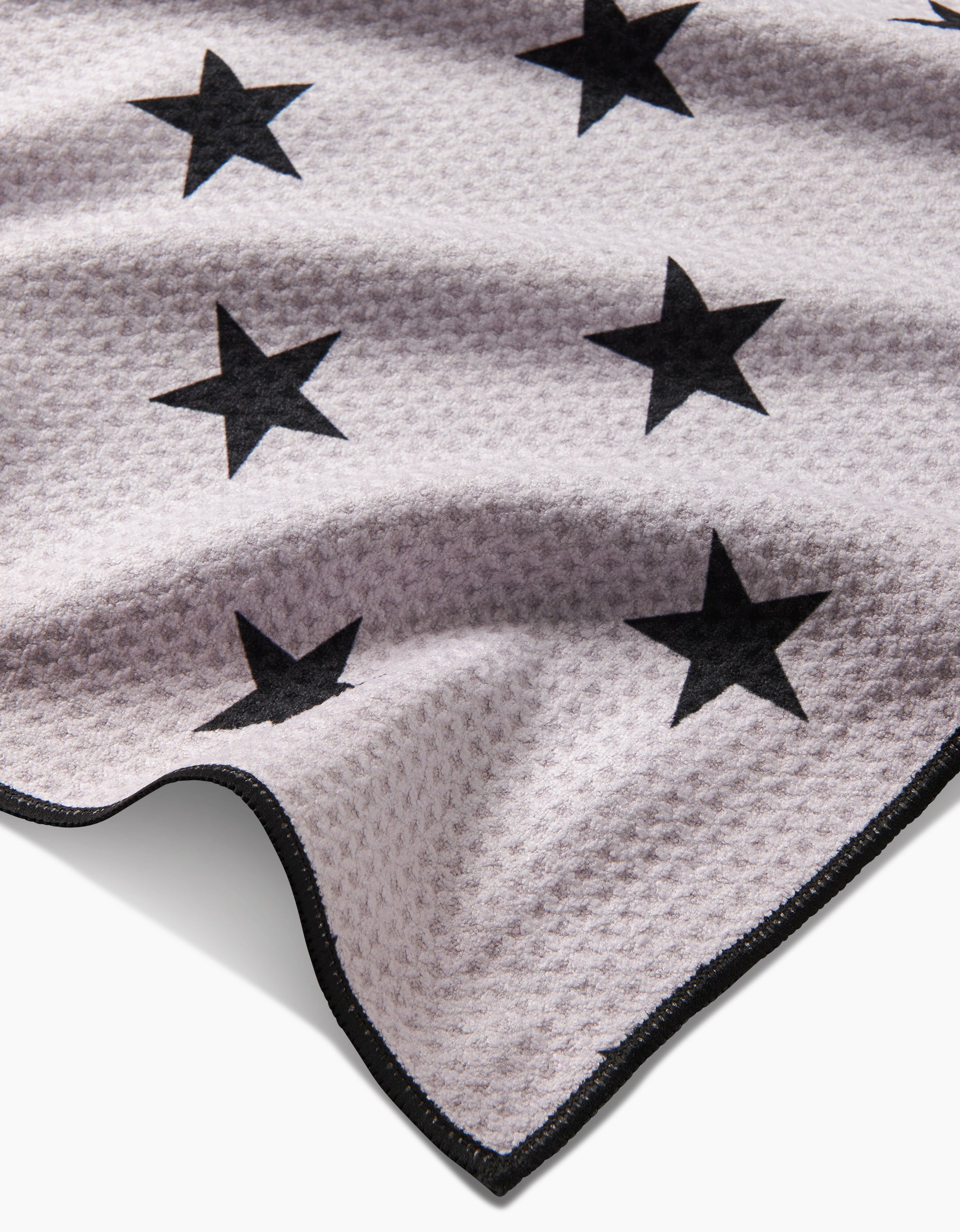 A close-up of Geometrys Bark, a light-toned, textured fabric made from recycled materials with black star patterns and a black-trimmed edge, shown lying flat with gentle folds.