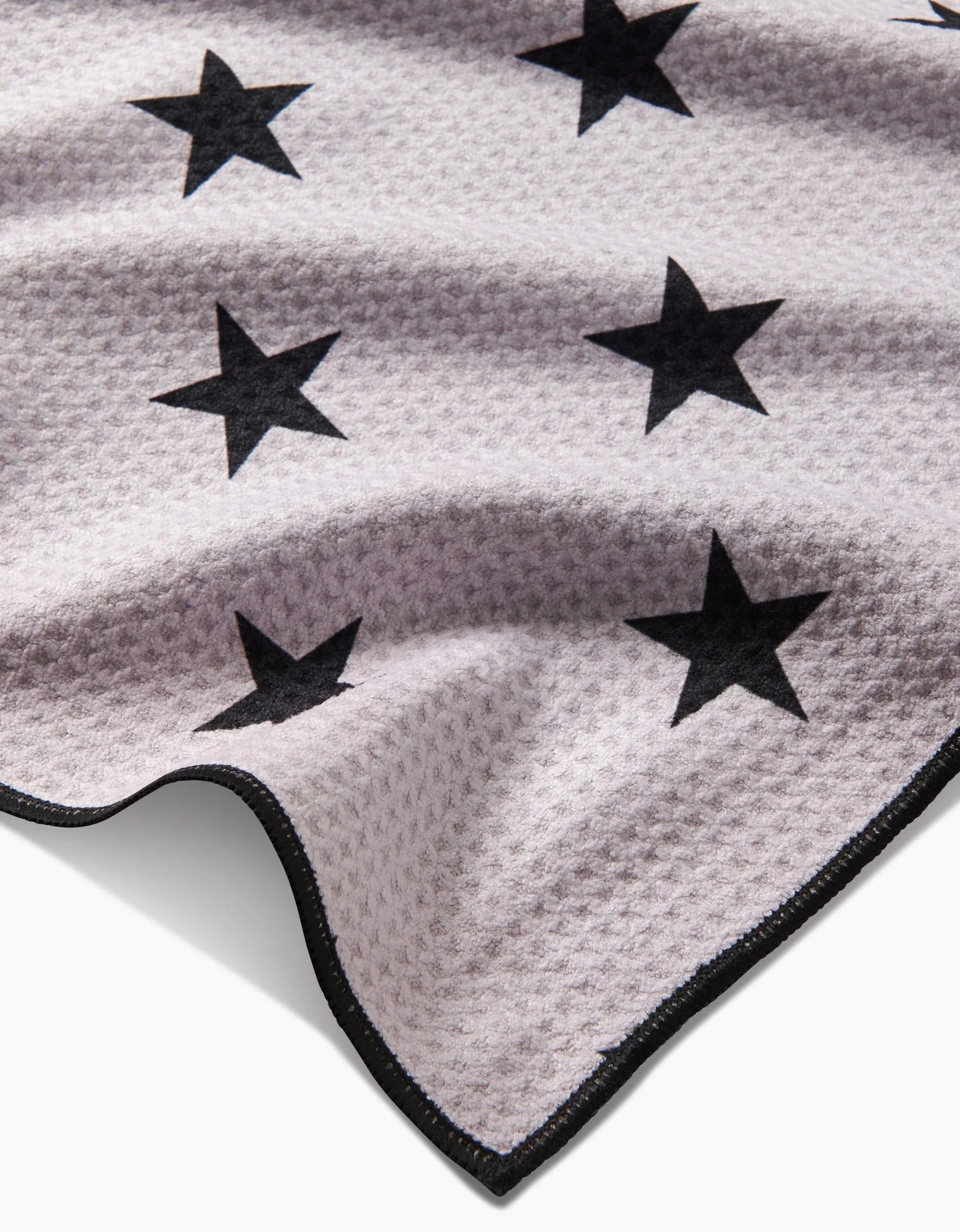 A close-up of Geometrys Bark, a light-toned, textured fabric made from recycled materials with black star patterns and a black-trimmed edge, shown lying flat with gentle folds.