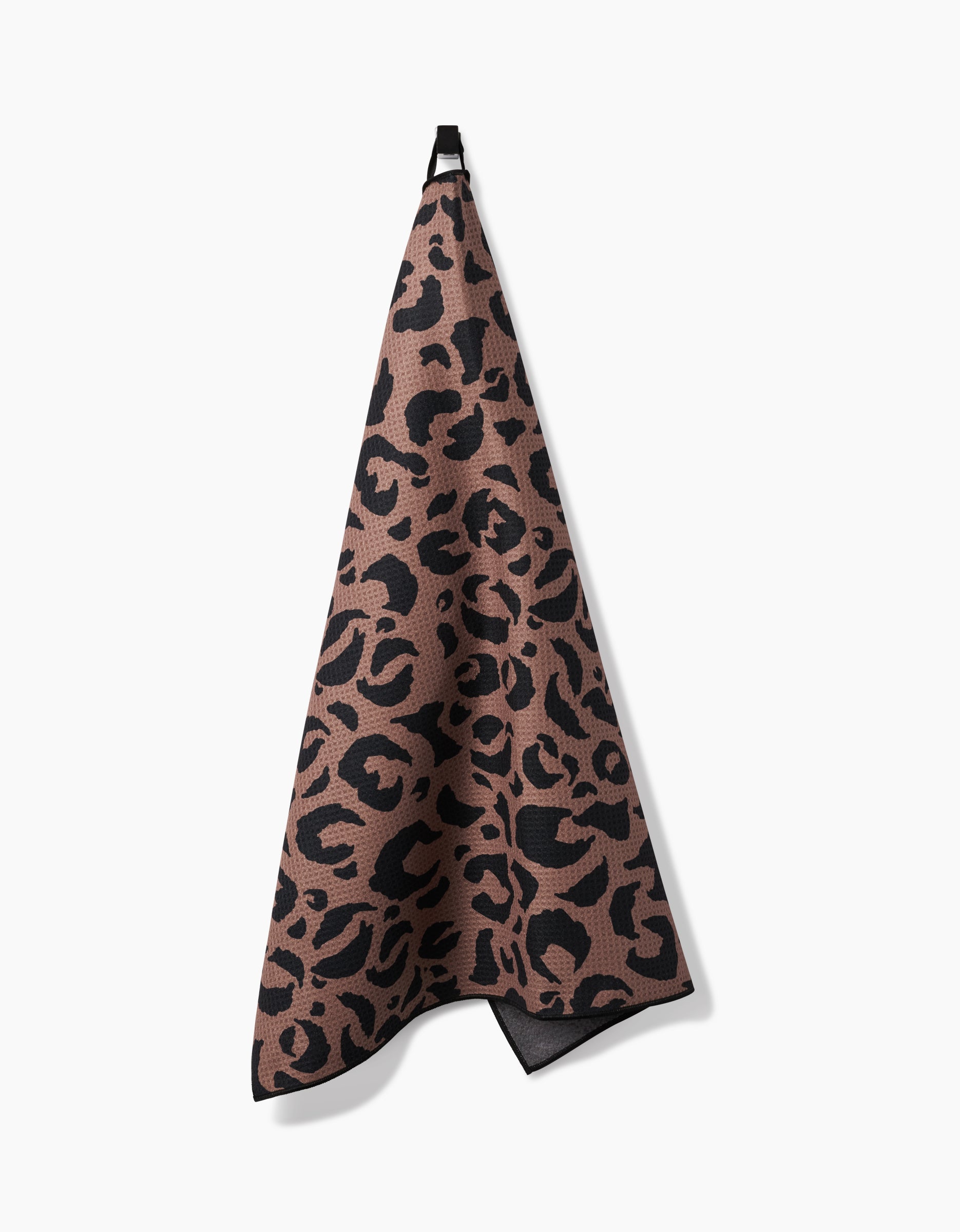 Geometry’s “Cheetah Hound” is a brown kitchen towel featuring a black leopard print, made from recycled materials, and hangs by a black loop against a white background.