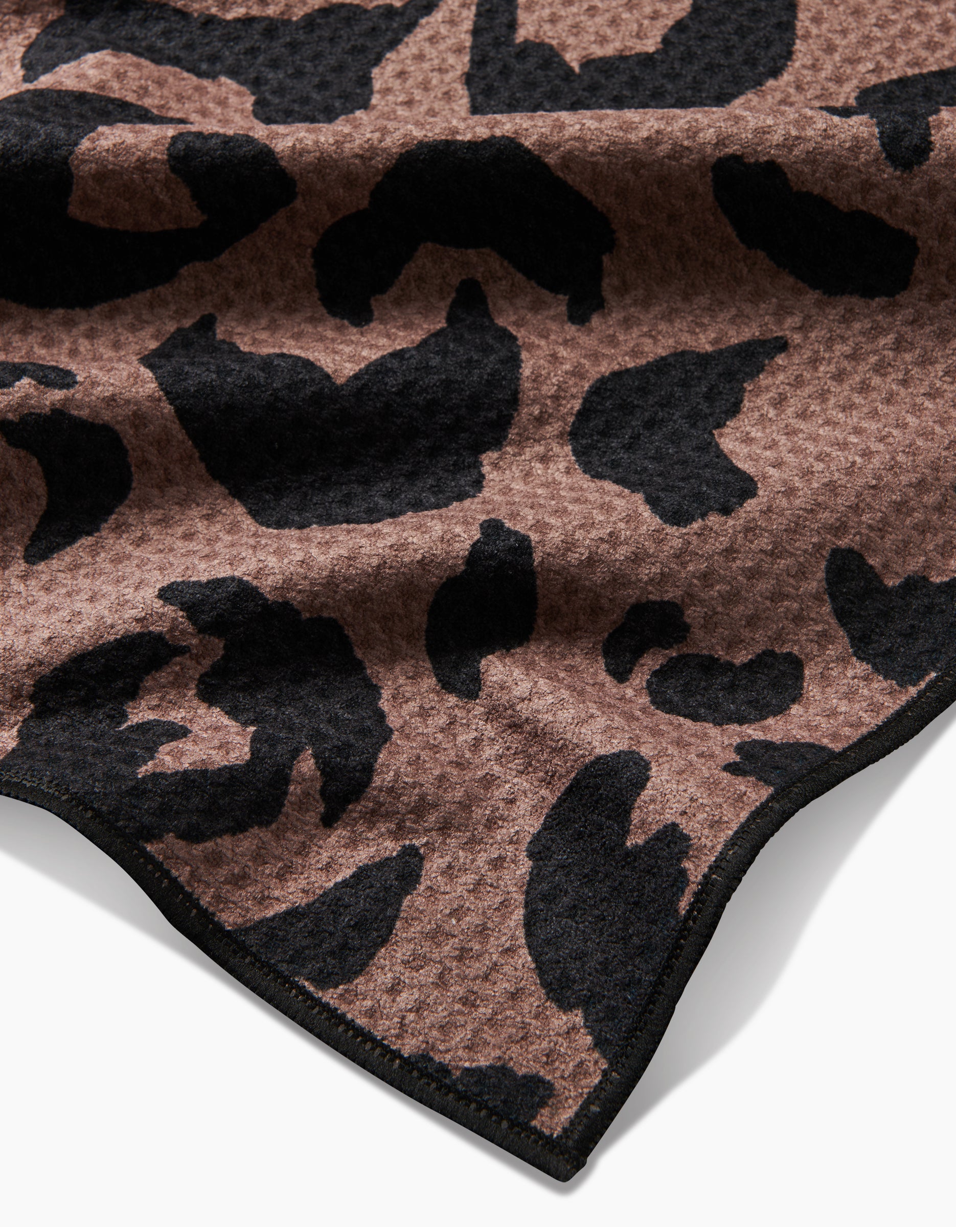 The Geometry Cheetah Hound dog towel is brown, ultra-absorbent, and made from recycled materials. It features a black cheetah print on a textured waffle weave with black-trimmed edges.