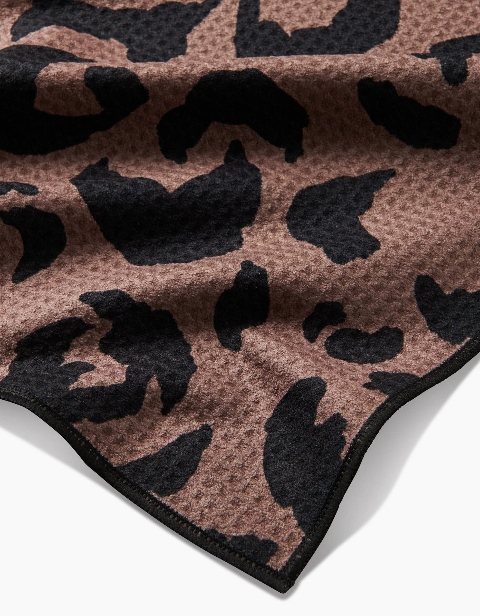 The Geometry Cheetah Hound dog towel is brown, ultra-absorbent, and made from recycled materials. It features a black cheetah print on a textured waffle weave with black-trimmed edges.