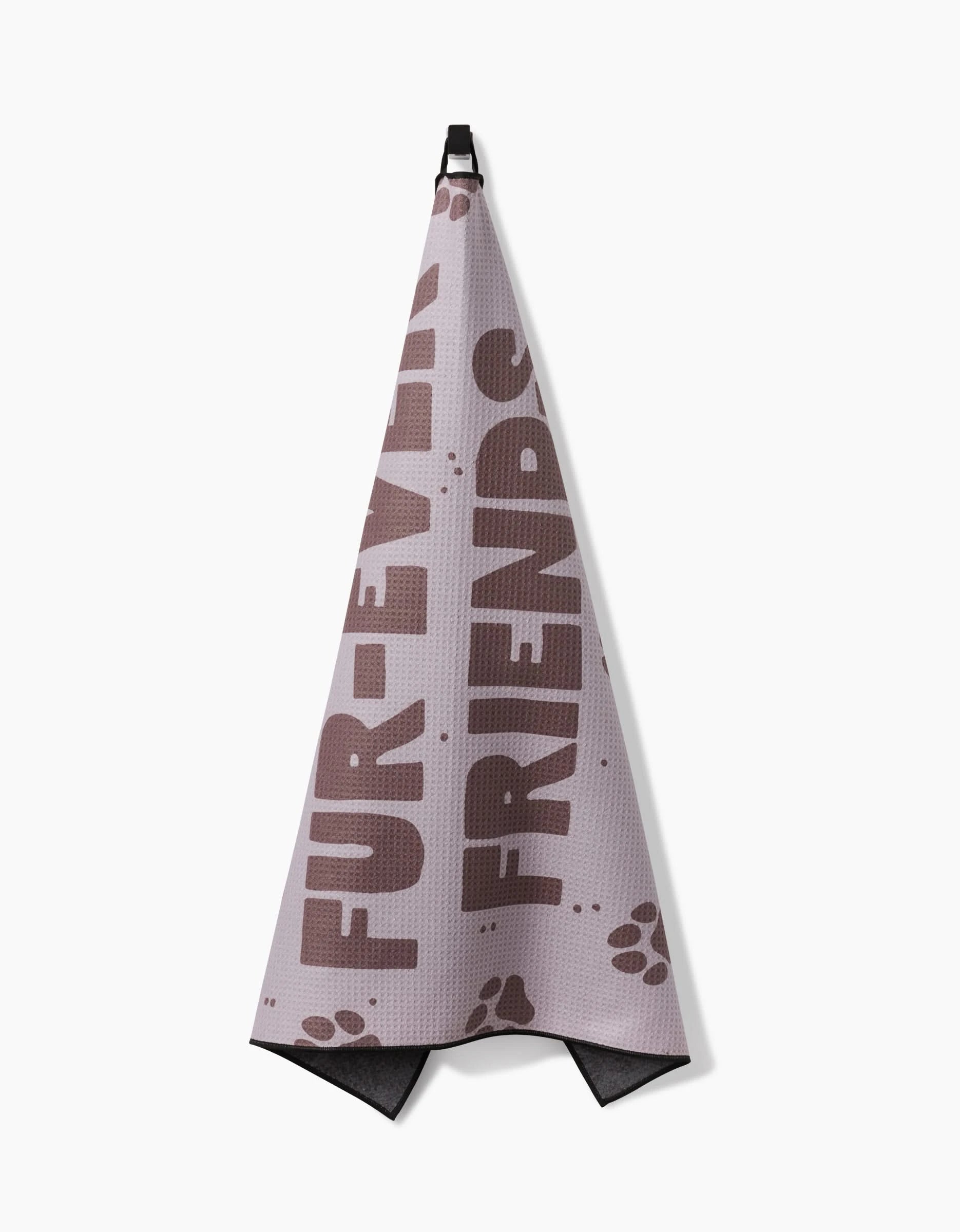The Geometry Fur Ever Friends pink dog towel, made from recycled materials, features brown FUR-EVER FRIENDS text and paw prints along the bottom. Its highly absorbent and hangs from a black hook.