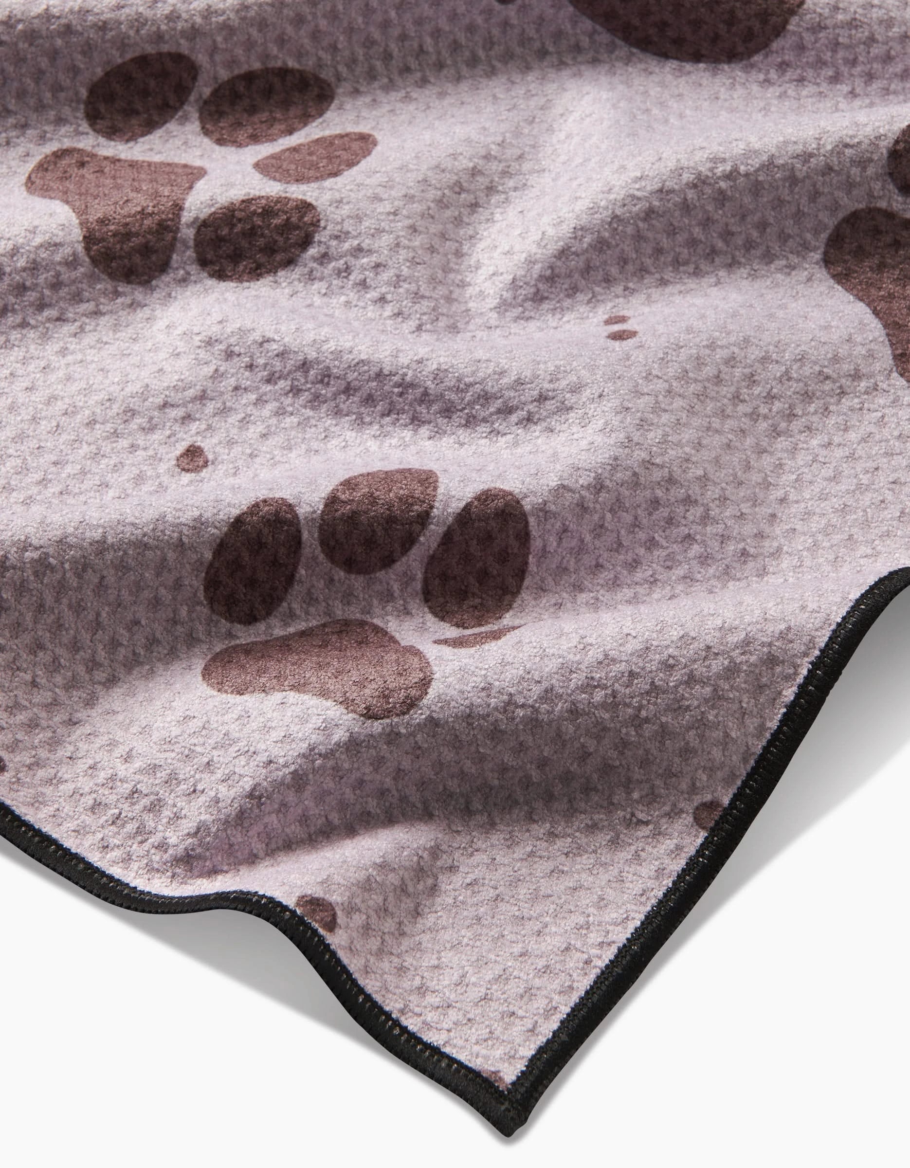 The Geometry Fur Ever Friends blanket is light purple with a textured surface, dark brown paw prints, and black trim. Made from recycled materials, it features gentle folds and creases for a cozy look.