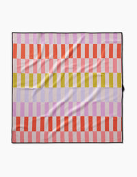 The Geometry Wag Wise blanket features a checkered pattern with horizontal stripes in red, light pink, yellow, and lavender, each formed by alternating squares. Made from recycled materials with a black border, it’s shown laid flat.