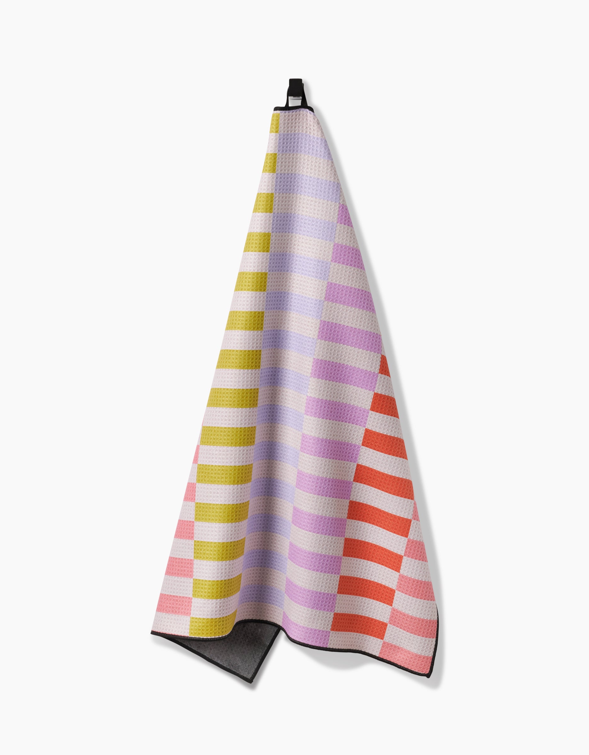 The Geometry Wag Wise towel, shown hanging from a black loop on a white backdrop, displays horizontal stripes in yellow, lavender, pink, and orange atop a textured pattern with a dark gray underside at the bottom corner.