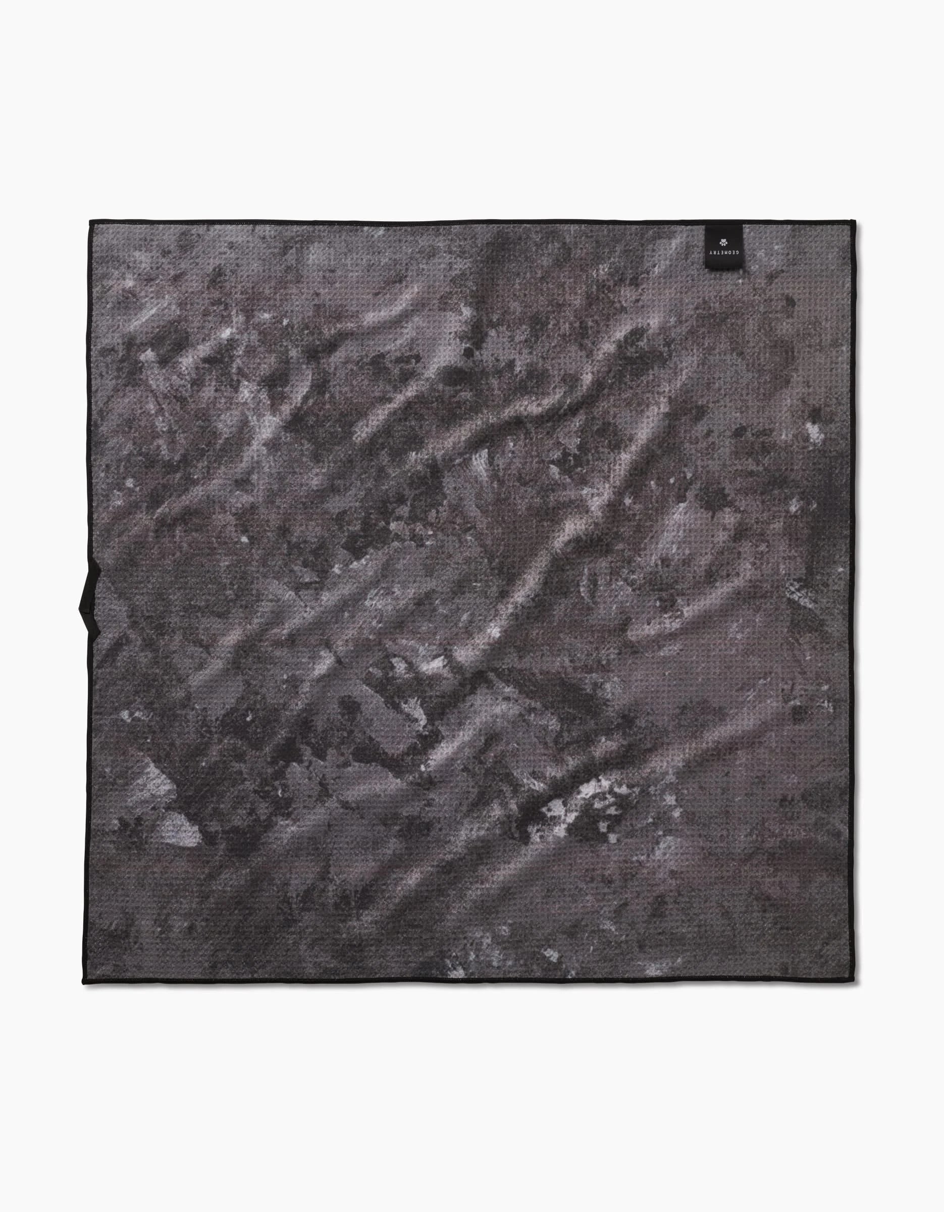 The Geometry Fur Ever Friends towel is a square, dark gray textured piece made from recycled materials, featuring black and light gray abstract patterns and a small black tag in one corner. Highly absorbent, it’s shown flat against a white background.