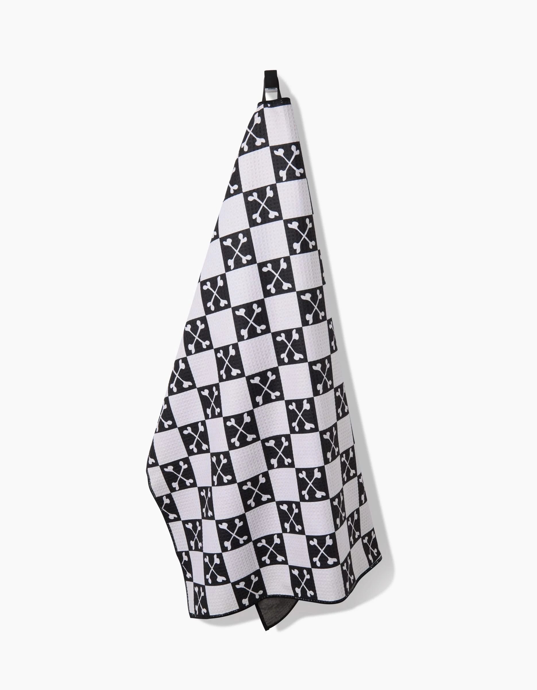 The Charlie Rowan Designs Boneyard Black Checkerboard dog towel hangs by a black loop, featuring black and white checks with white intersecting arrow logos. Made from recycled materials, its stylish and highly absorbent.