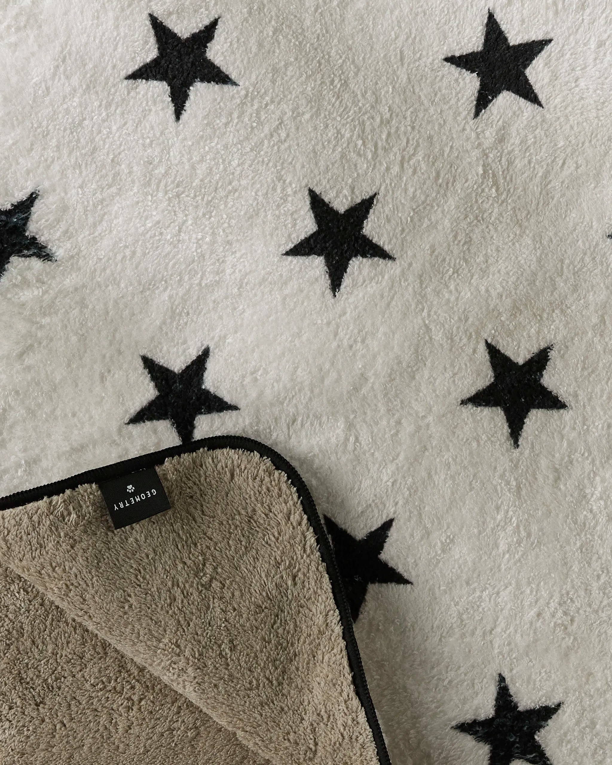 A close-up of Geometry's Bark dog towel, featuring black star patterns. A corner is flipped to reveal its tan, fuzzy recycled underside and a small black tag with white text.