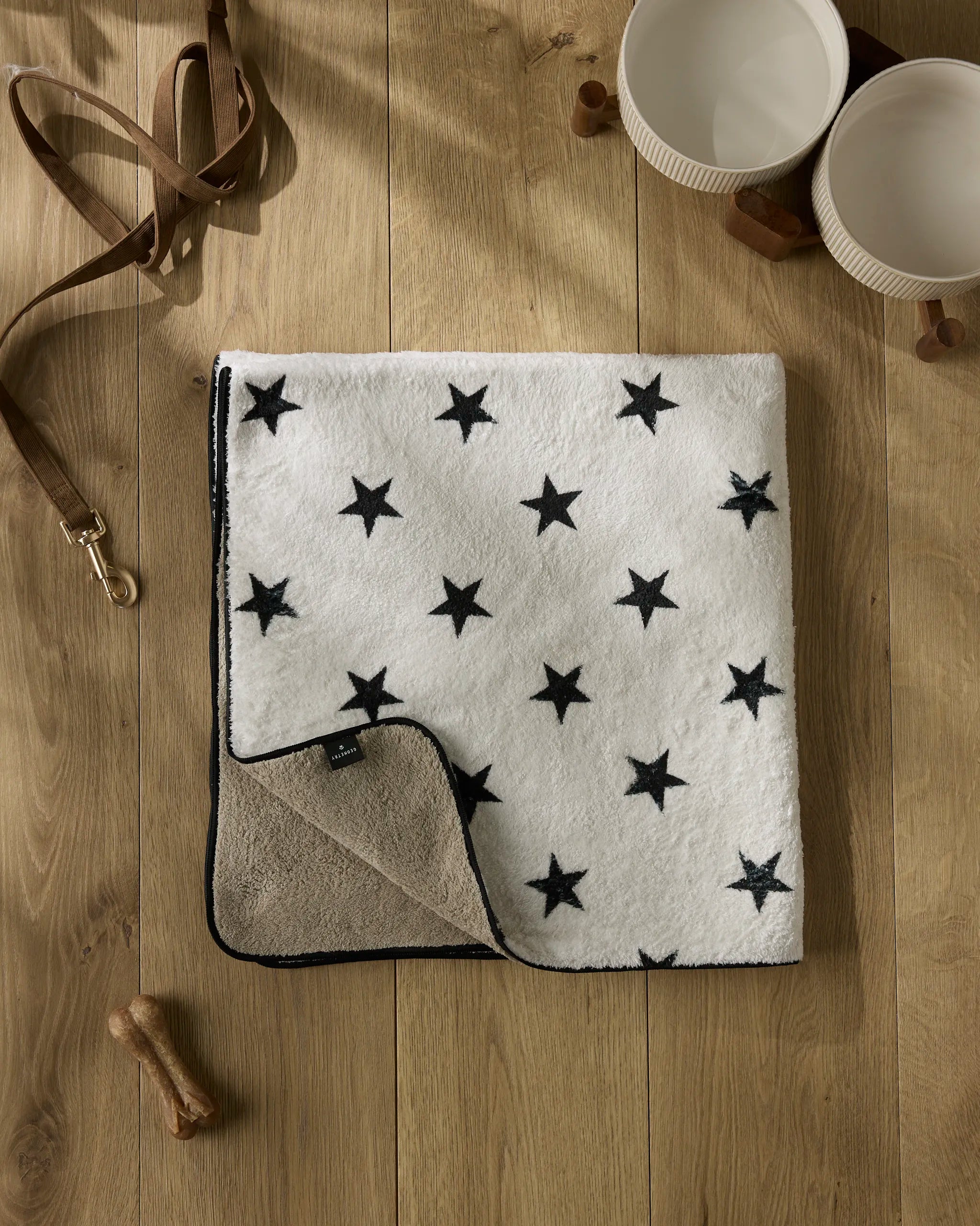 The Geometry Bark dog towel, crafted from recycled materials in white and beige with black stars, rests on a wooden floor beside two white bowls, a brown leather leash, and a dog bone.