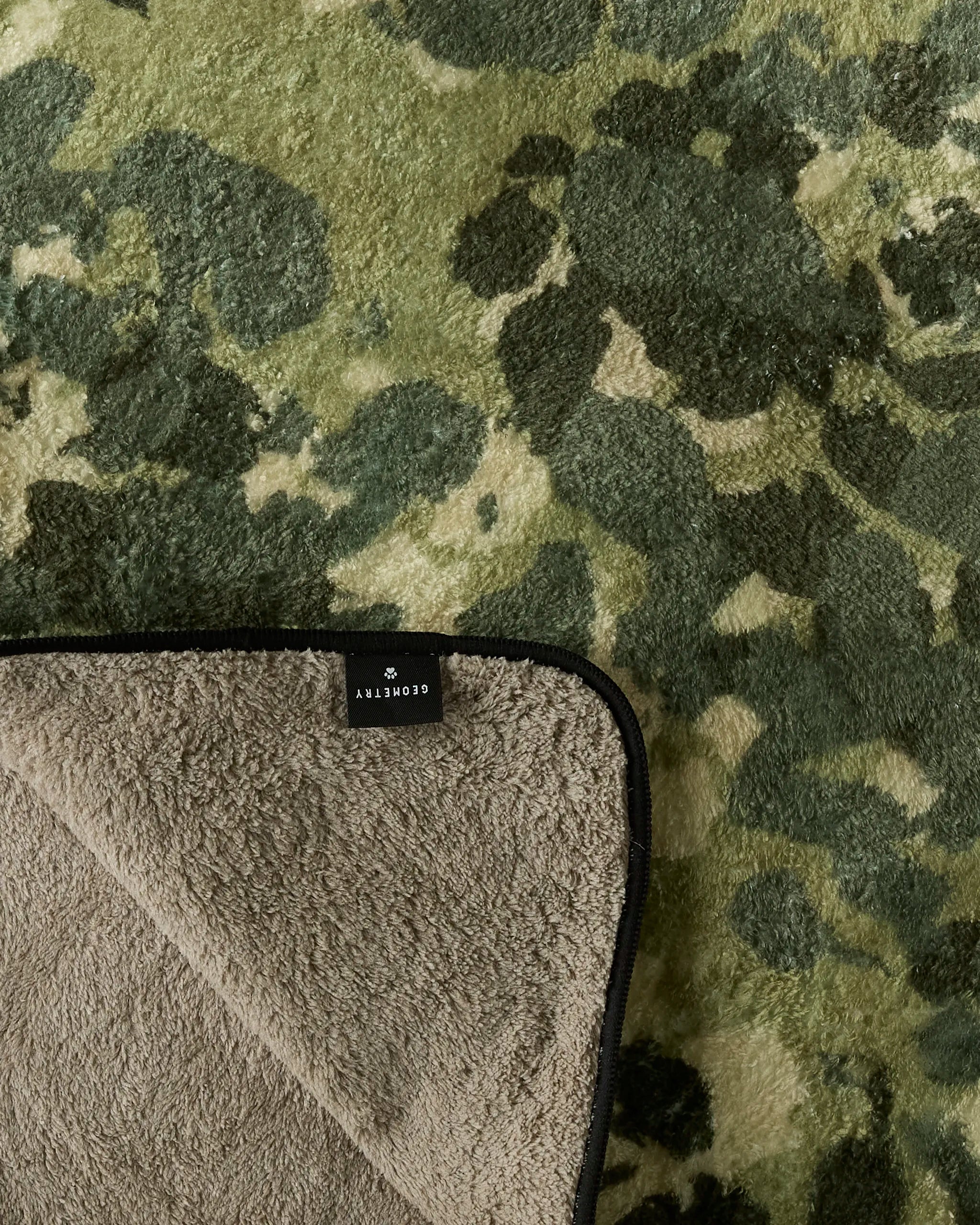 A close-up of Geometry’s "Camo" green and beige camouflage rug with one corner flipped to show its ultra-absorbent dog towel underside and a black label reading “Geometry.”.