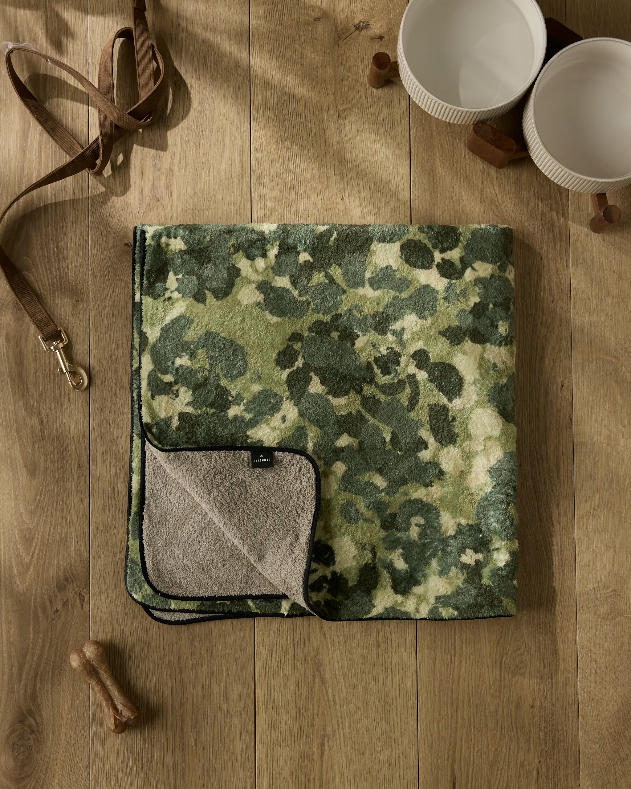 The Geometry Camo dog towel, featuring a green camouflage pattern and soft gray underside, is folded on a wooden floor with two white bowls, a brown leather leash, and two dog treats nearby.
