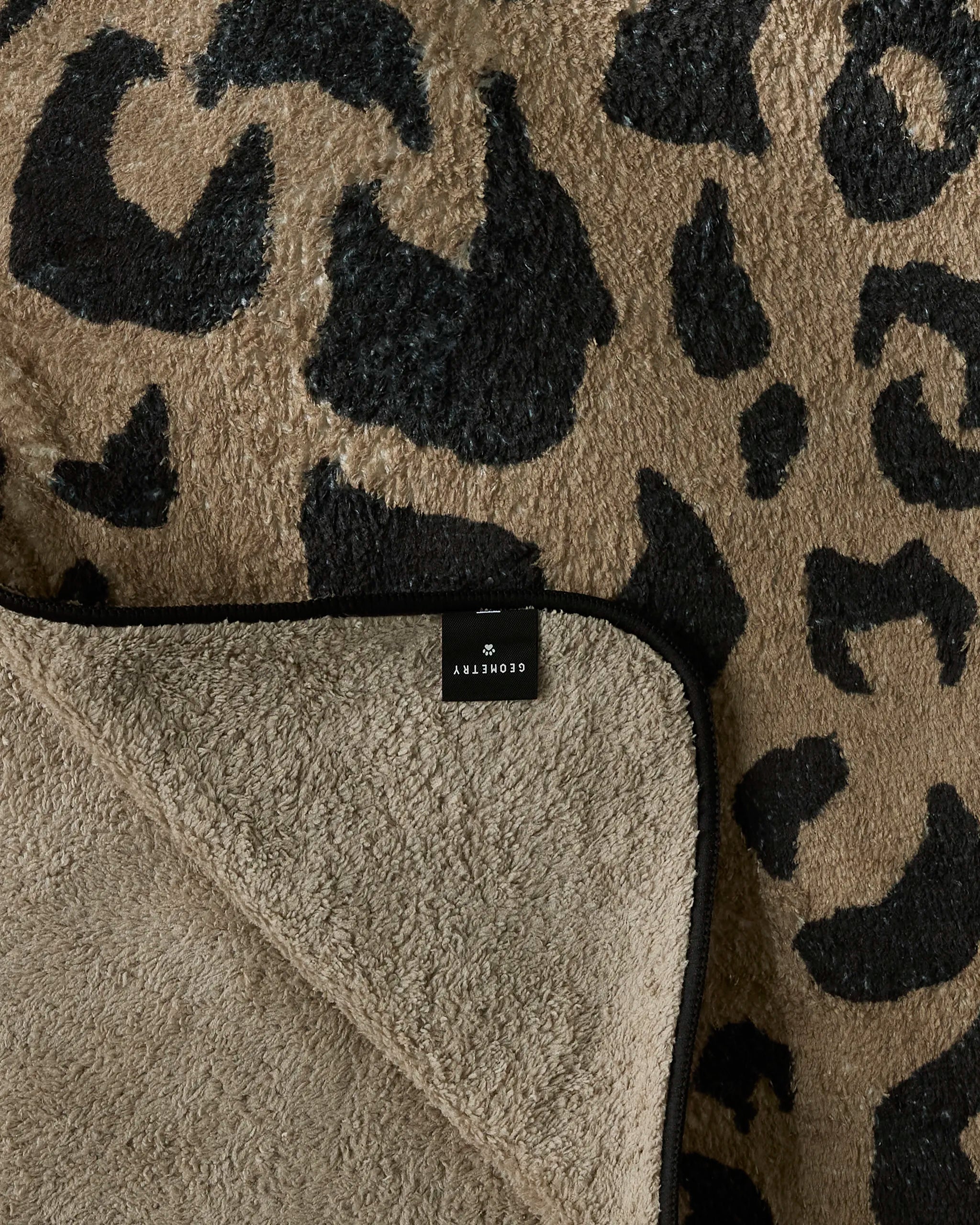 The Geometry Cheetah Hound is a plush dog towel with a black and tan leopard print, partially folded to reveal its plain tan underside and a label reading "Geometry.