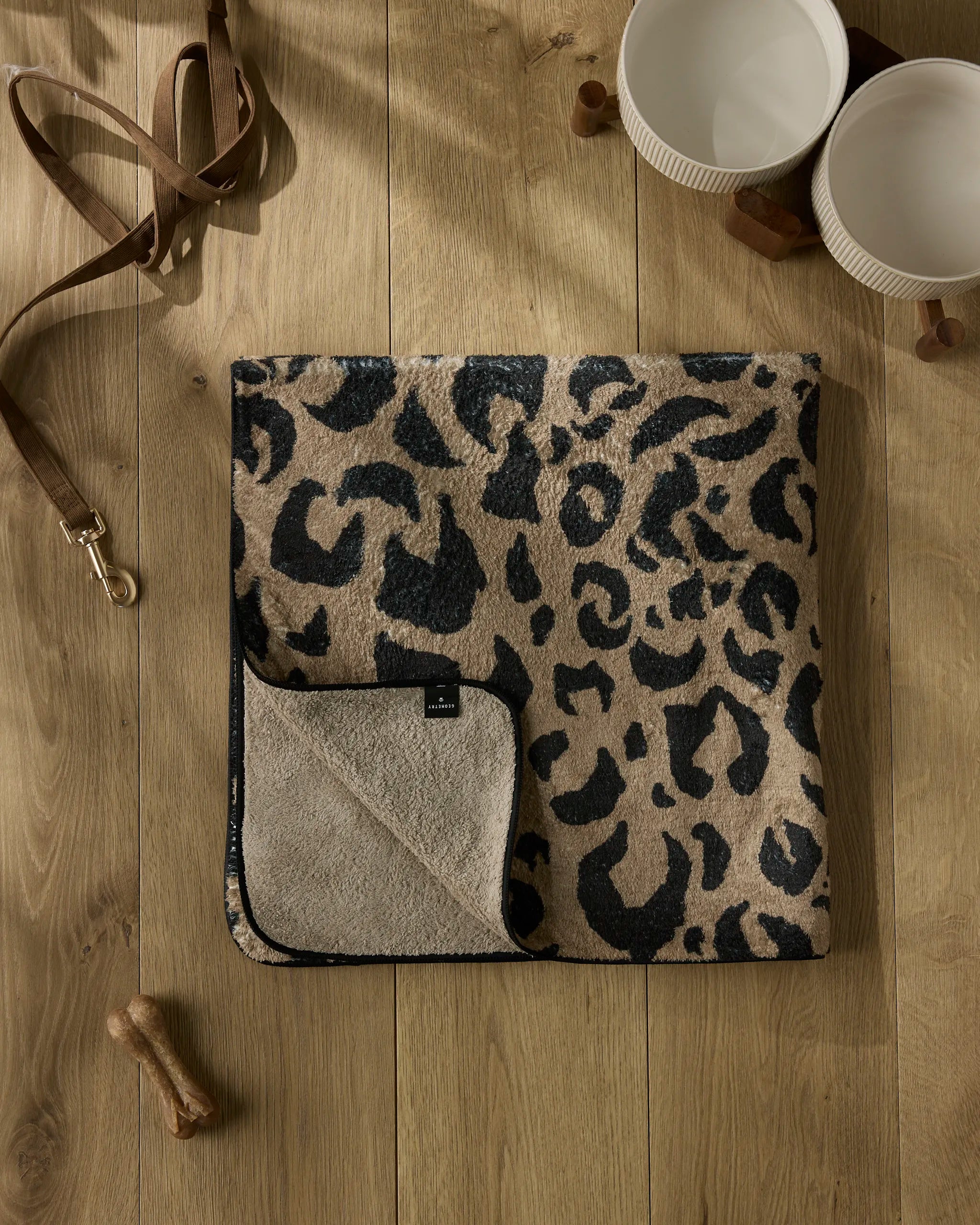 The Geometry Cheetah Hound towel, ultra-absorbent and folded with a cheetah print and beige underside, rests on a wooden floor beside two white bowls, a brown dog leash, a wooden bone toy, and a dog treat.