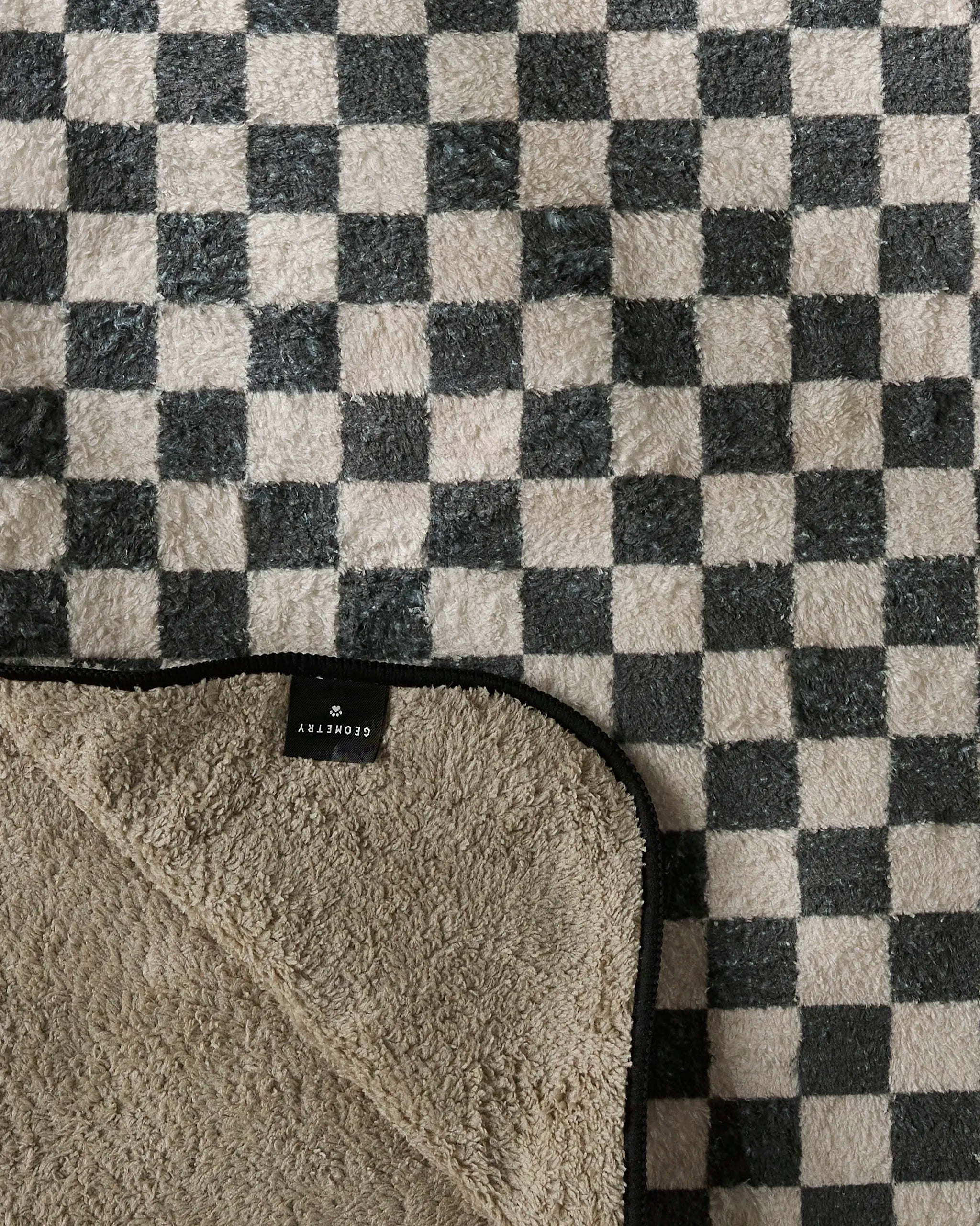 A soft, checkered blanket with a black-and-white grid pattern. One corner is flipped to reveal a beige underside and a black label reading Geometry—crafted as the ultra-absorbent Cool Pup dog towel from recycled materials.