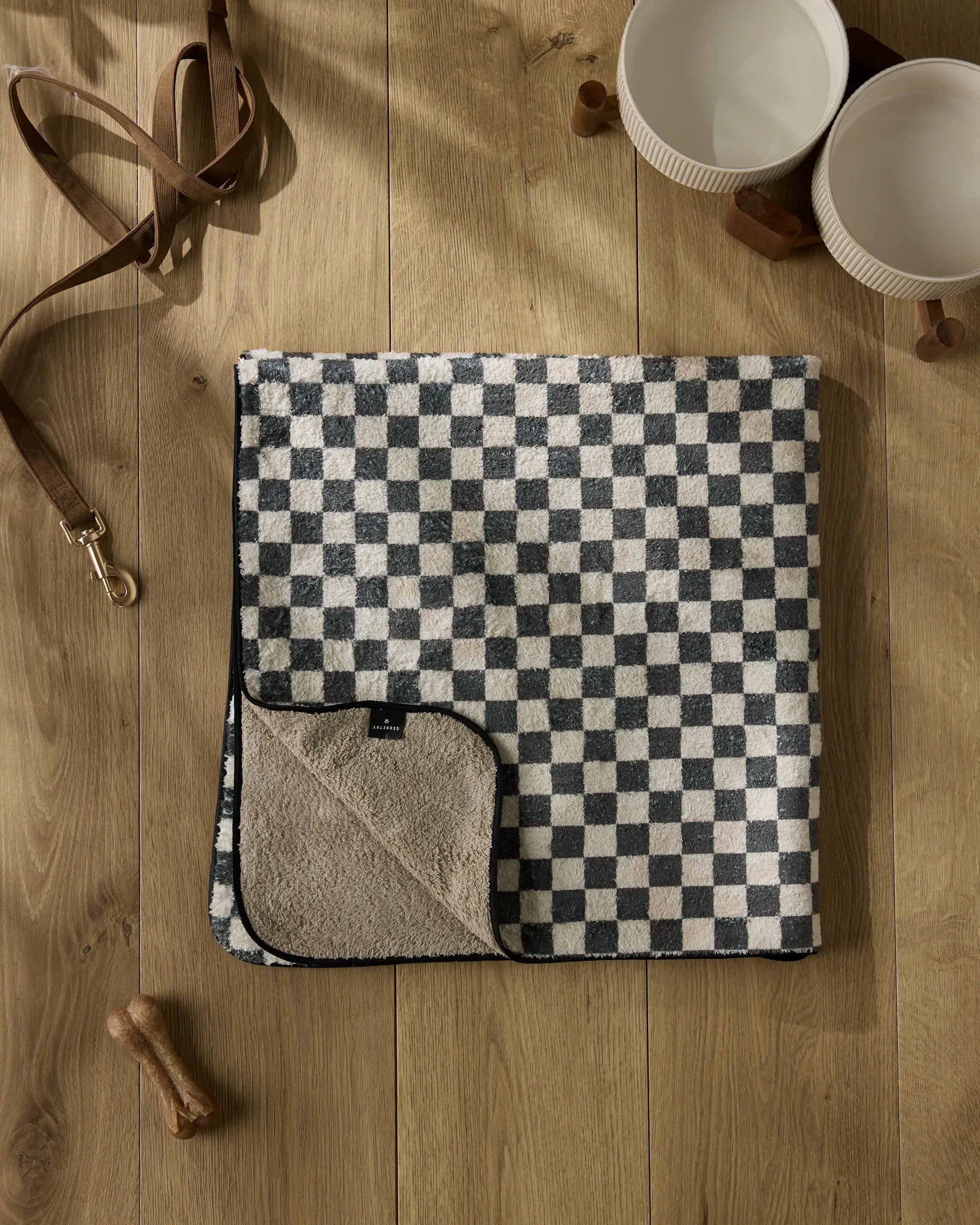 A folded Cool Pup dog towel by Geometry, featuring black and beige squares, rests on a wooden floor with a brown leash, two white bowls, a bone-shaped treat, and two wooden dog bowl stands nearby.