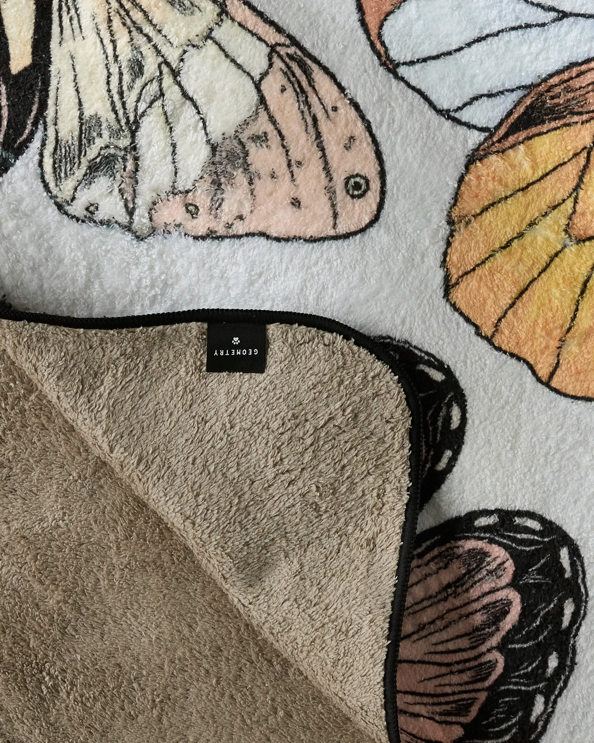 A close-up of the Geometry "Cute Butterflies" ultra-absorbent dog towel, partially folded to reveal its soft beige underside, with detailed butterfly wings in beige, orange, and black.