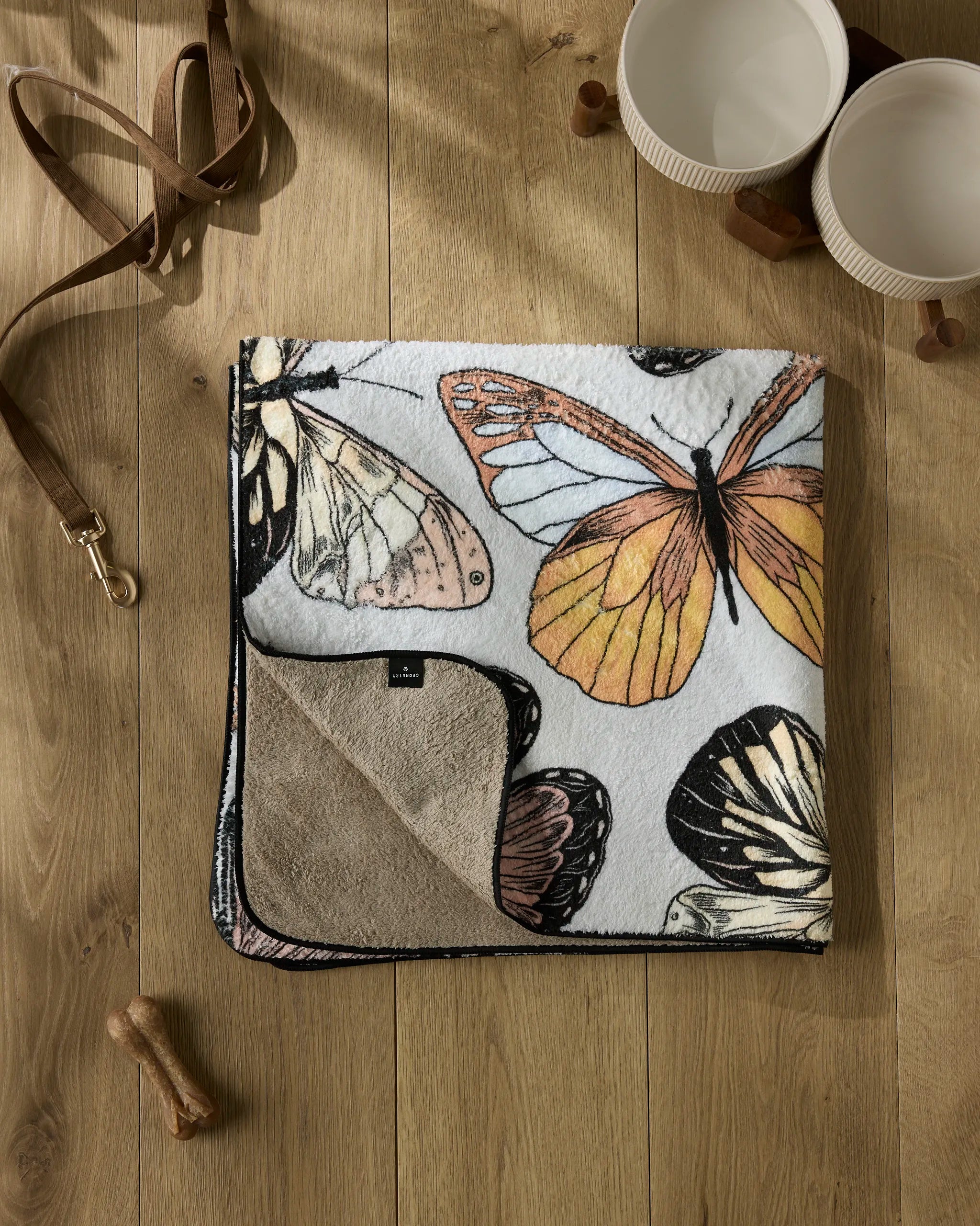 The Geometry 'Cute Butterflies' plush dog towel is folded on a wooden floor with its soft beige underside showing. Nearby are two white bowls, a brown leash, wooden blocks, and a small dog bone.