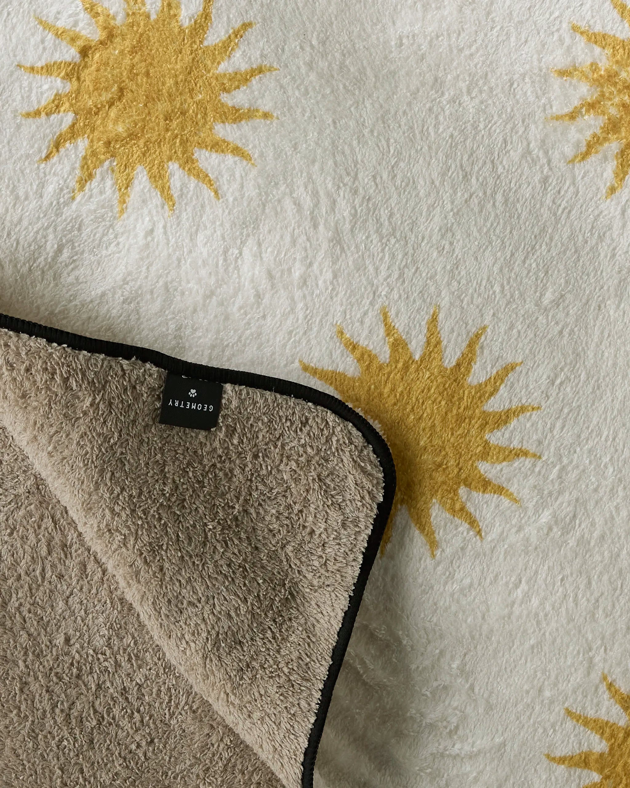 A soft, fluffy white Geometry dog towel called "Feeling Sunny" features yellow sunburst patterns, lies flat with one corner flipped to show a beige underside and a black CLOSETRTY label.