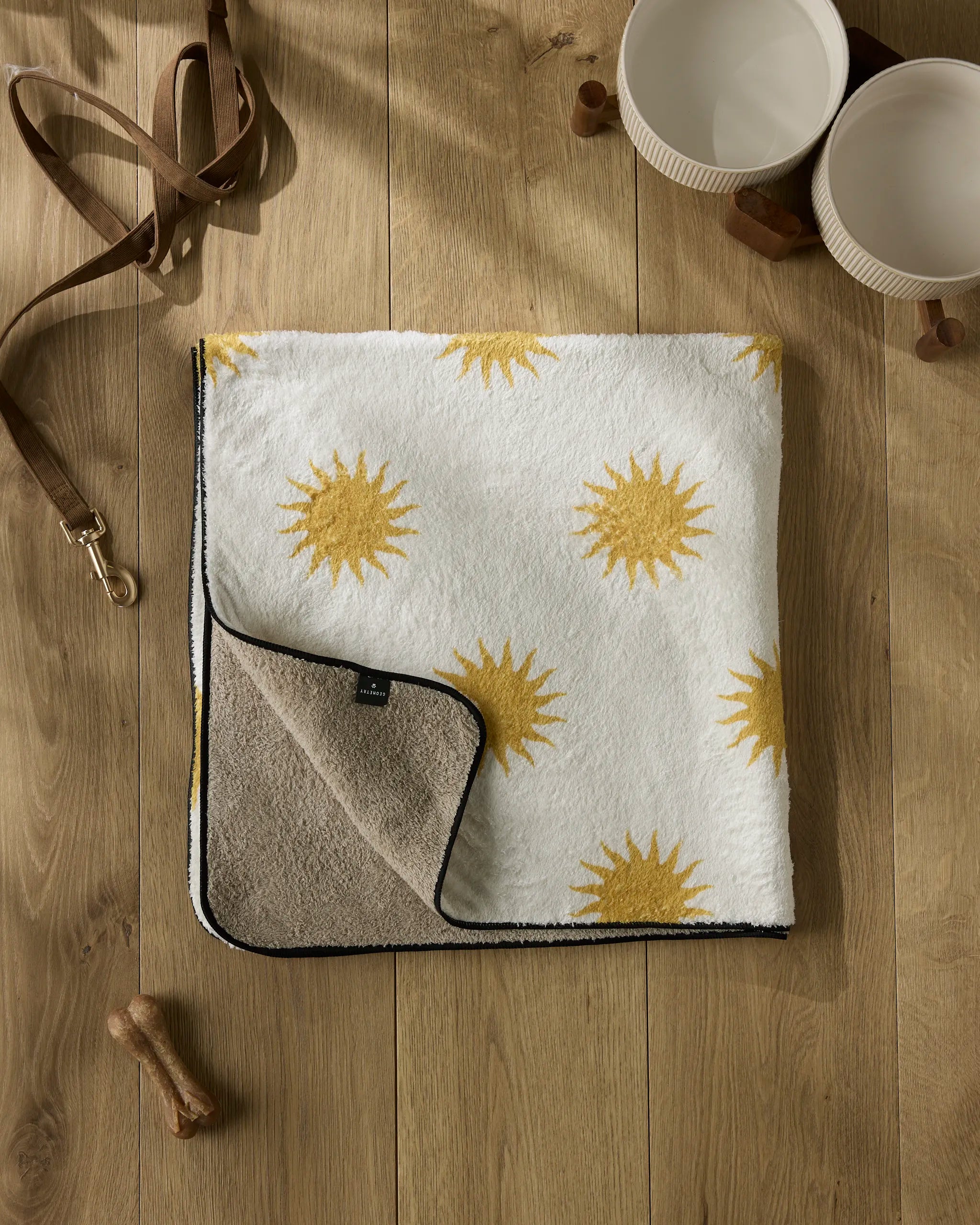The Geometry Feeling Sunny plush dog towel with yellow sun patterns lies folded on a wooden floor, alongside two cream pet bowls, a brown dog leash, and some treats. The towel’s underside is beige.