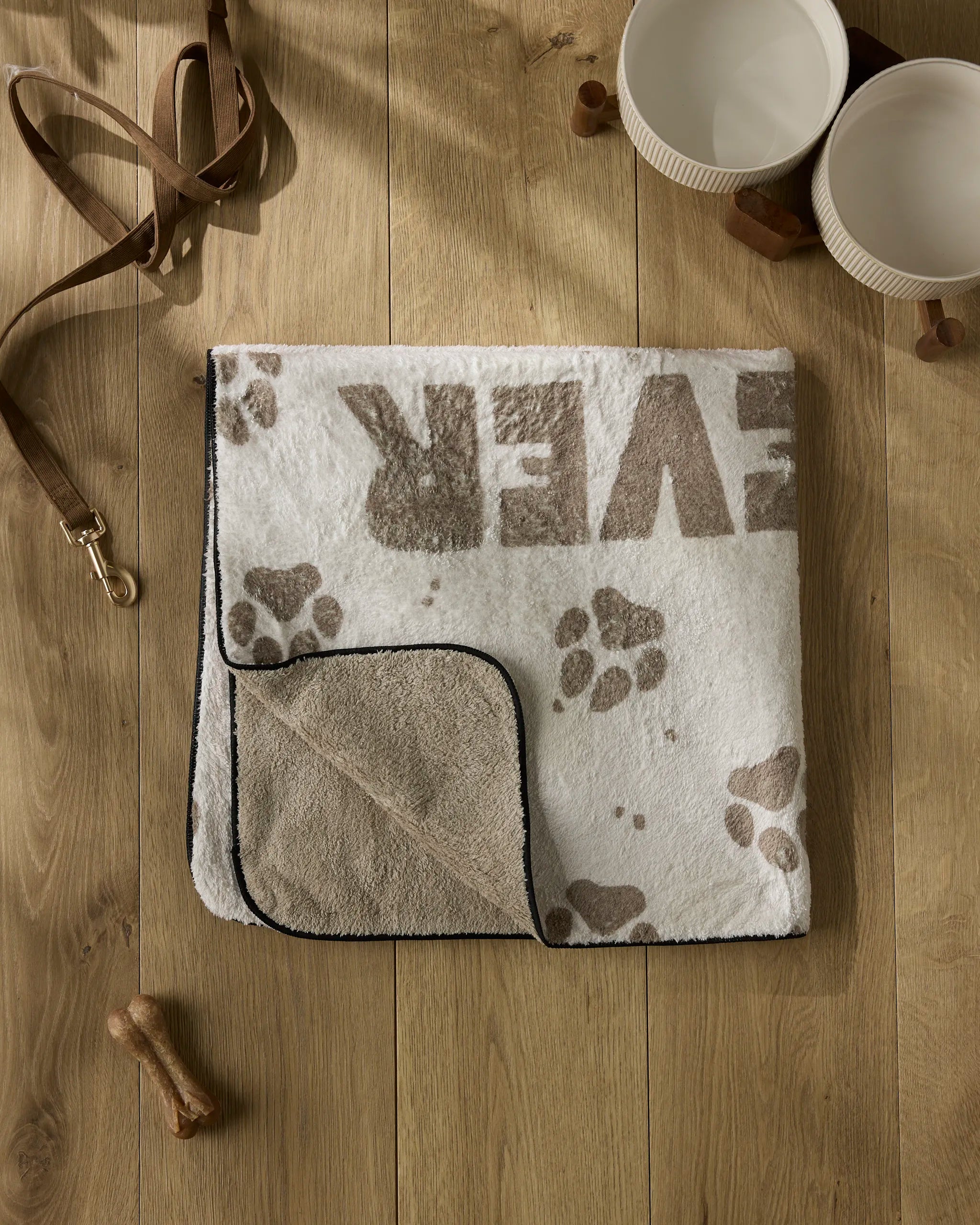 A folded Geometry Fur Ever Friends ultra-absorbent dog blanket with paw prints and "Fur Ever Friends" text, surrounded by a leash, two bowls, and a dog bone on a wooden floor.