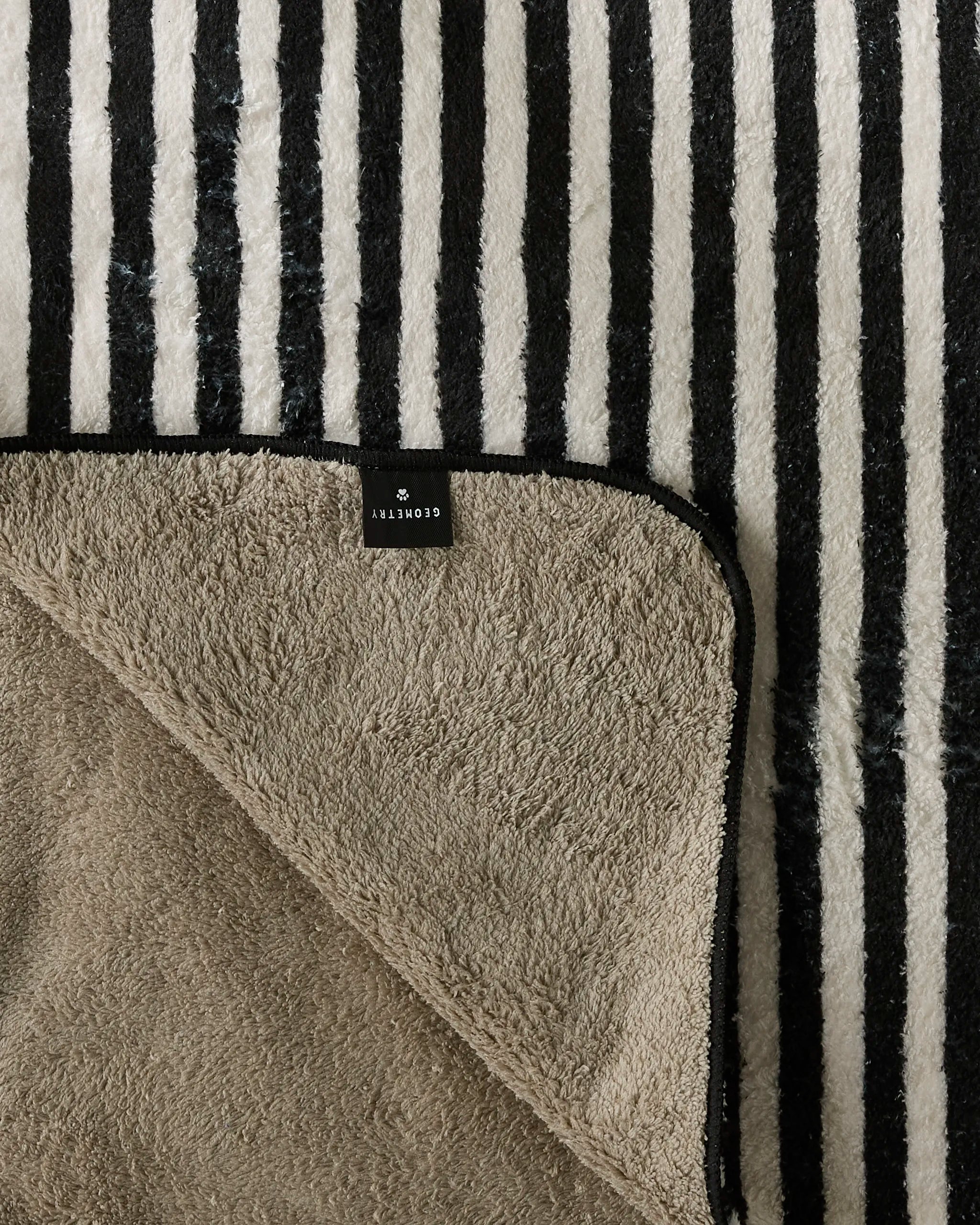 A soft, textured Good Pup dog towel by Geometry with black and white stripes on one side and plain beige on the other, partially folded to reveal both designs. A black tag labeled Geometry is visible on the striped side.
