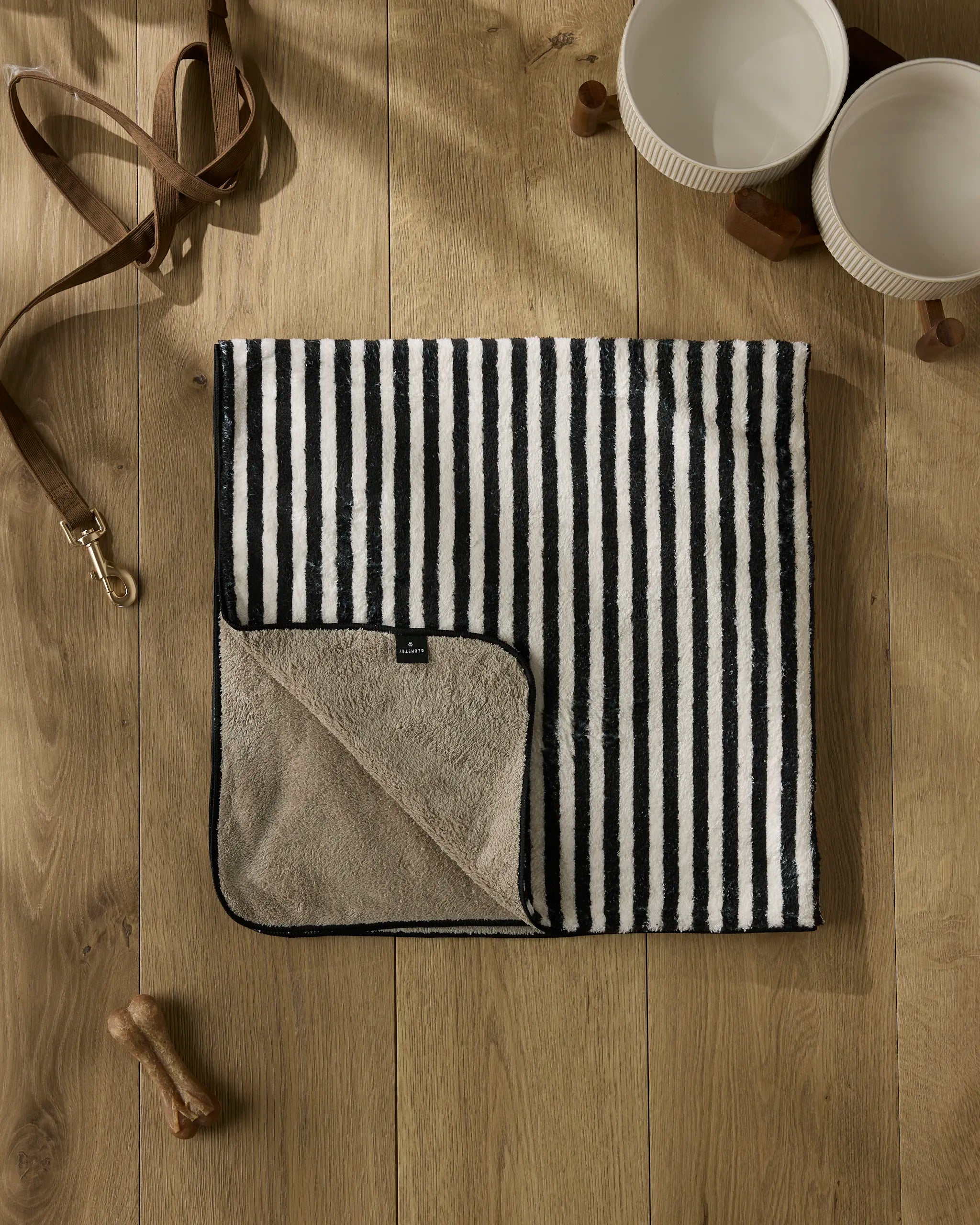 The Geometry Good Pup ultra-absorbent dog towel in black and white stripes with a beige underside is laid flat on a wooden floor, surrounded by two dog bowls, a leather leash, a bone-shaped treat, and a collar.