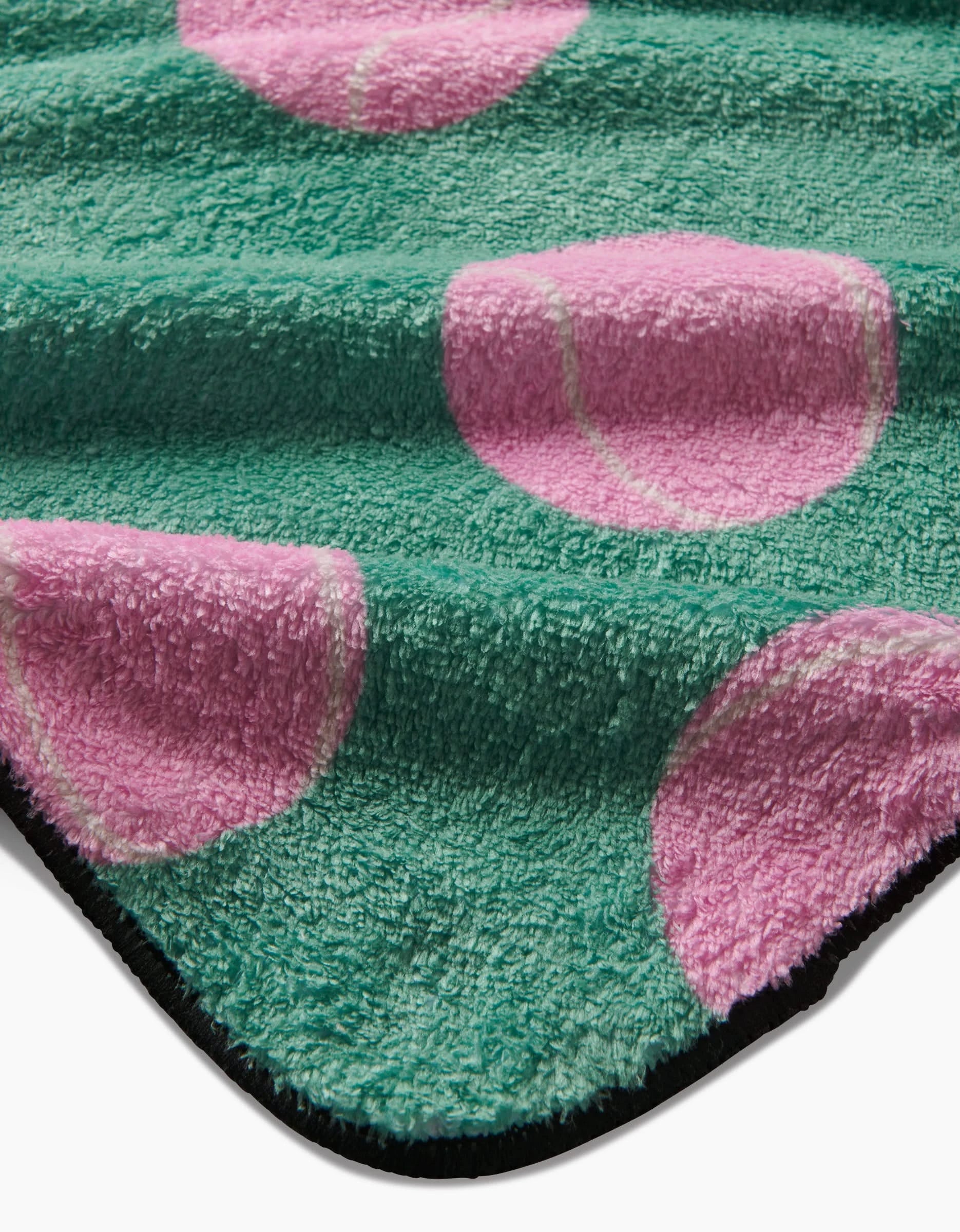 A close-up of Geometrys Pink Ball dog towel: a soft, ultra-absorbent towel made from recycled materials with a textured green fabric, large pink tennis ball patterns, and a black edge.