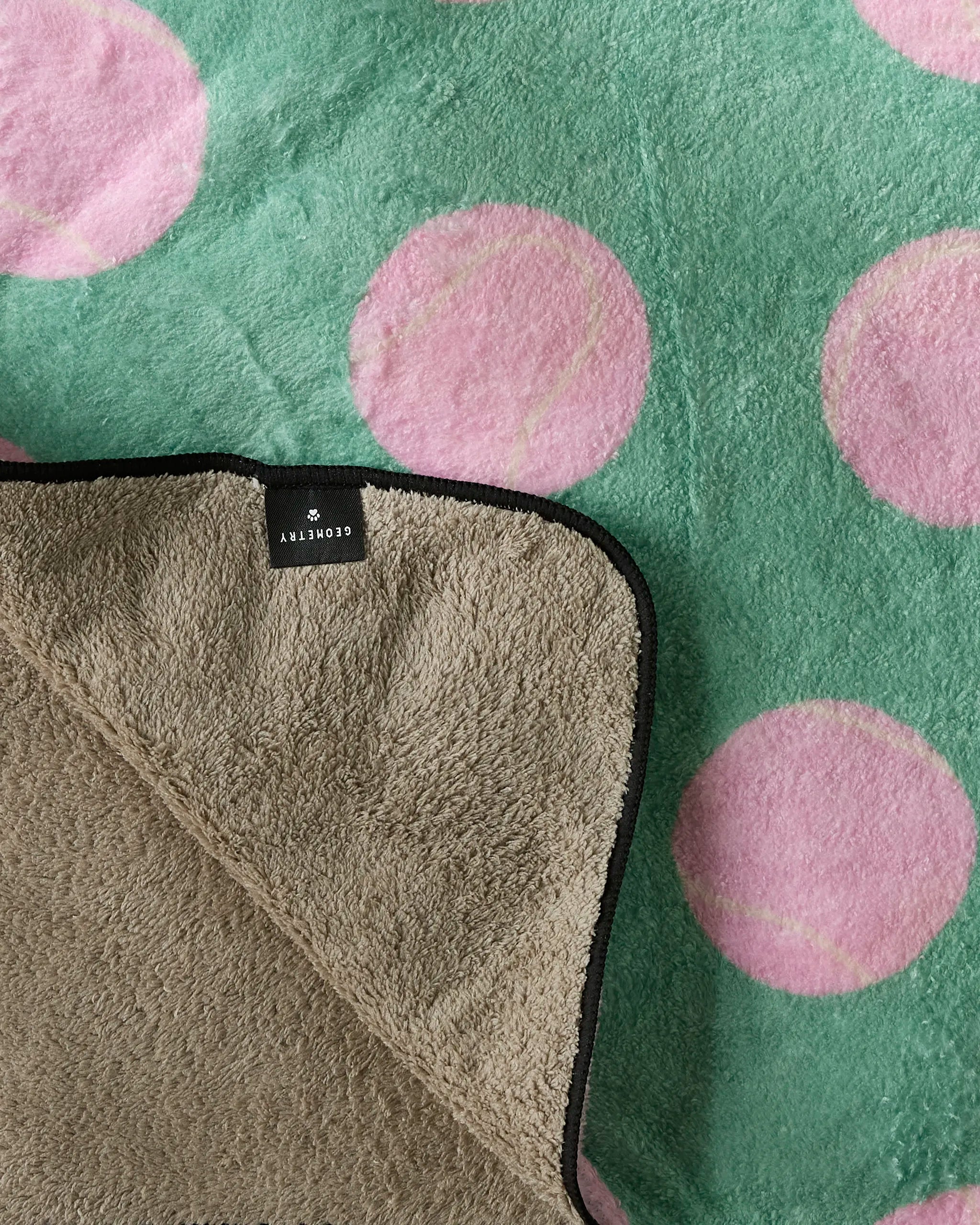 A close-up of Geometry's Pink Ball green blanket, patterned with pink tennis balls. An ultra-absorbent dog towel with a black edge and a STORYBNY label rests on top.