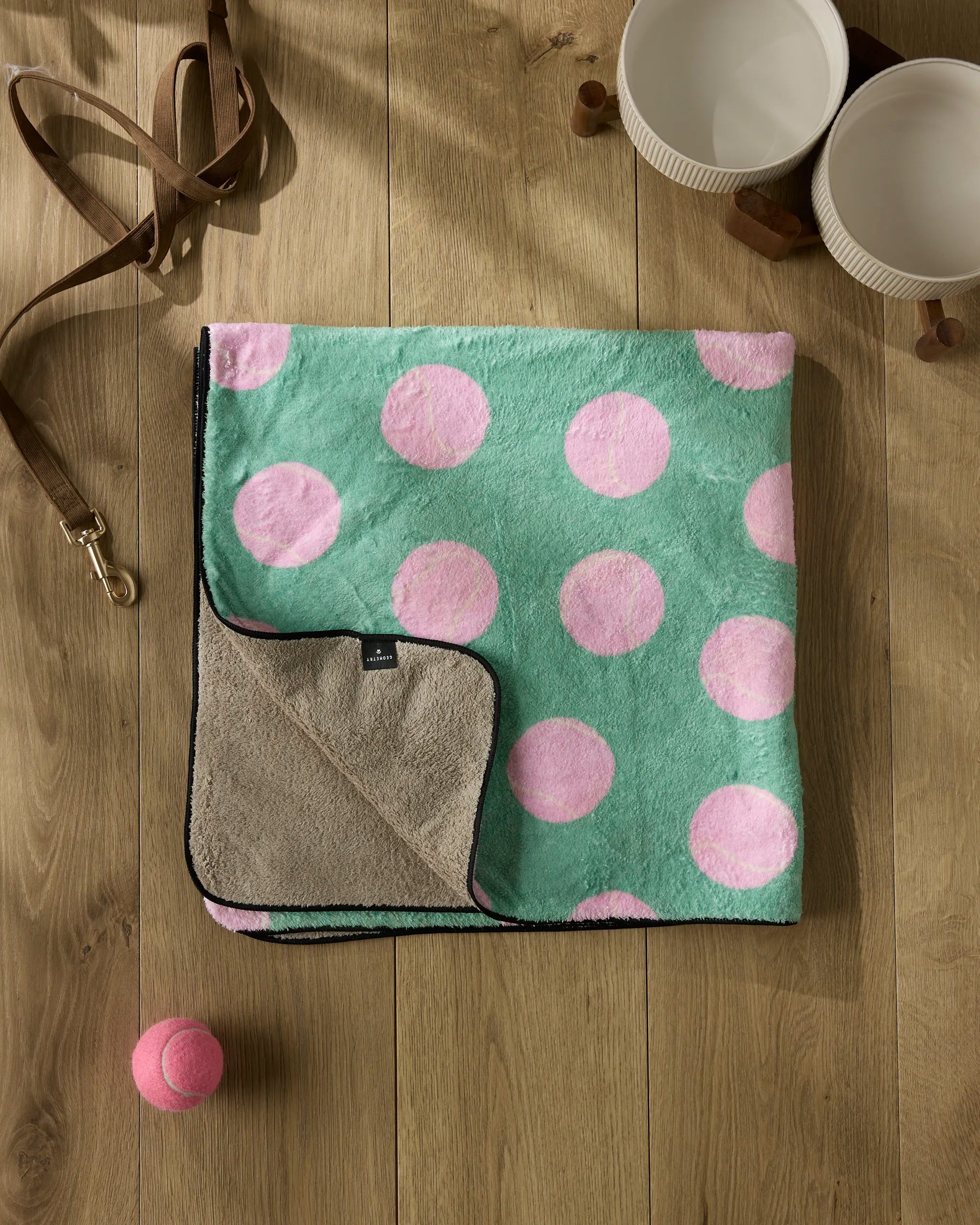 A plush dog towel with large pink polka dots is neatly folded on a wooden floor beside Geometry's Pink Ball, a brown leash, two white bowls, and several brown chew toys.