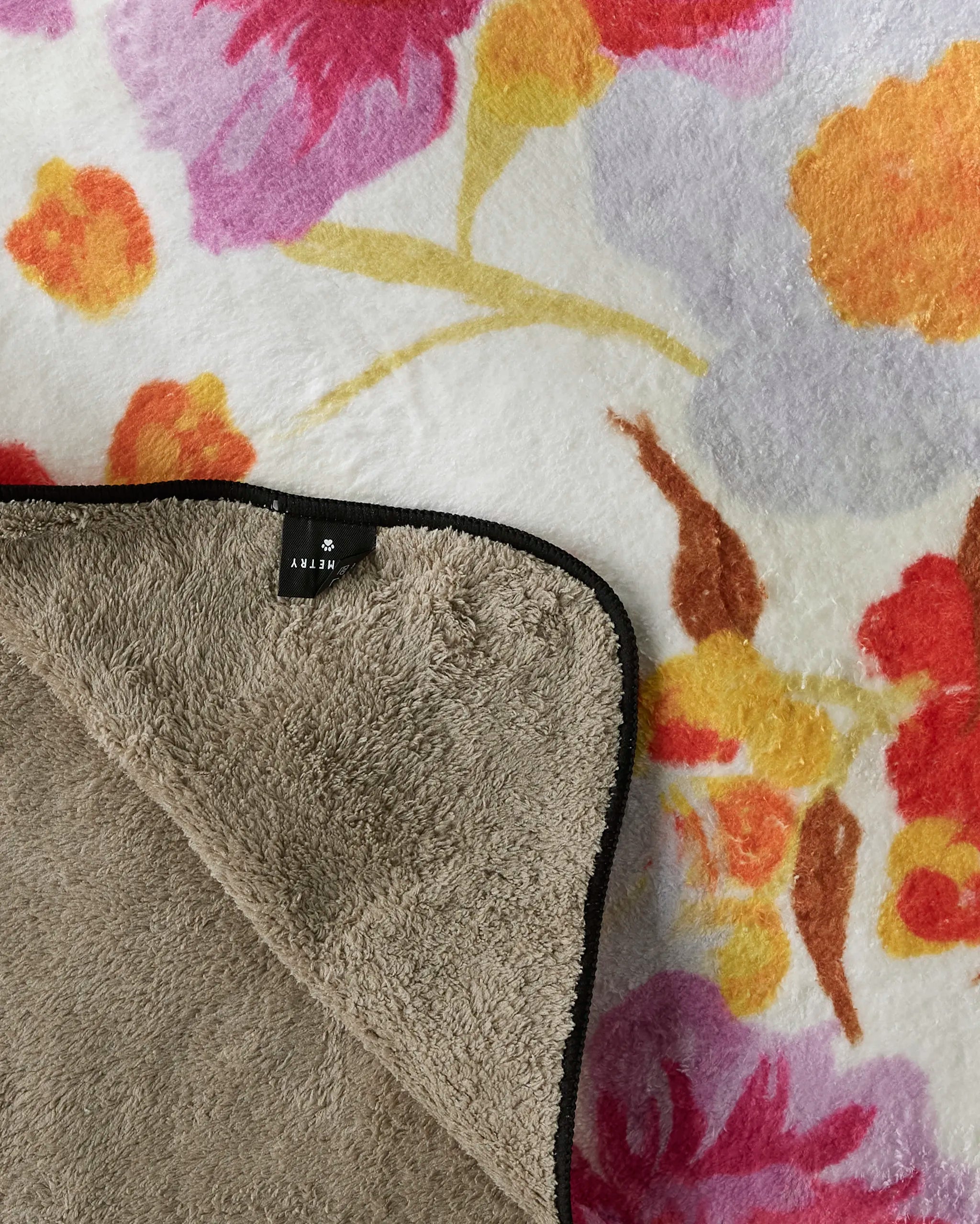 A close-up of Geometry's Pup Florals dog towel shows a plush, soft texture with vibrant red, orange, pink, and yellow floral patterns. A folded corner reveals the plain beige underside and a black “S” size tag.