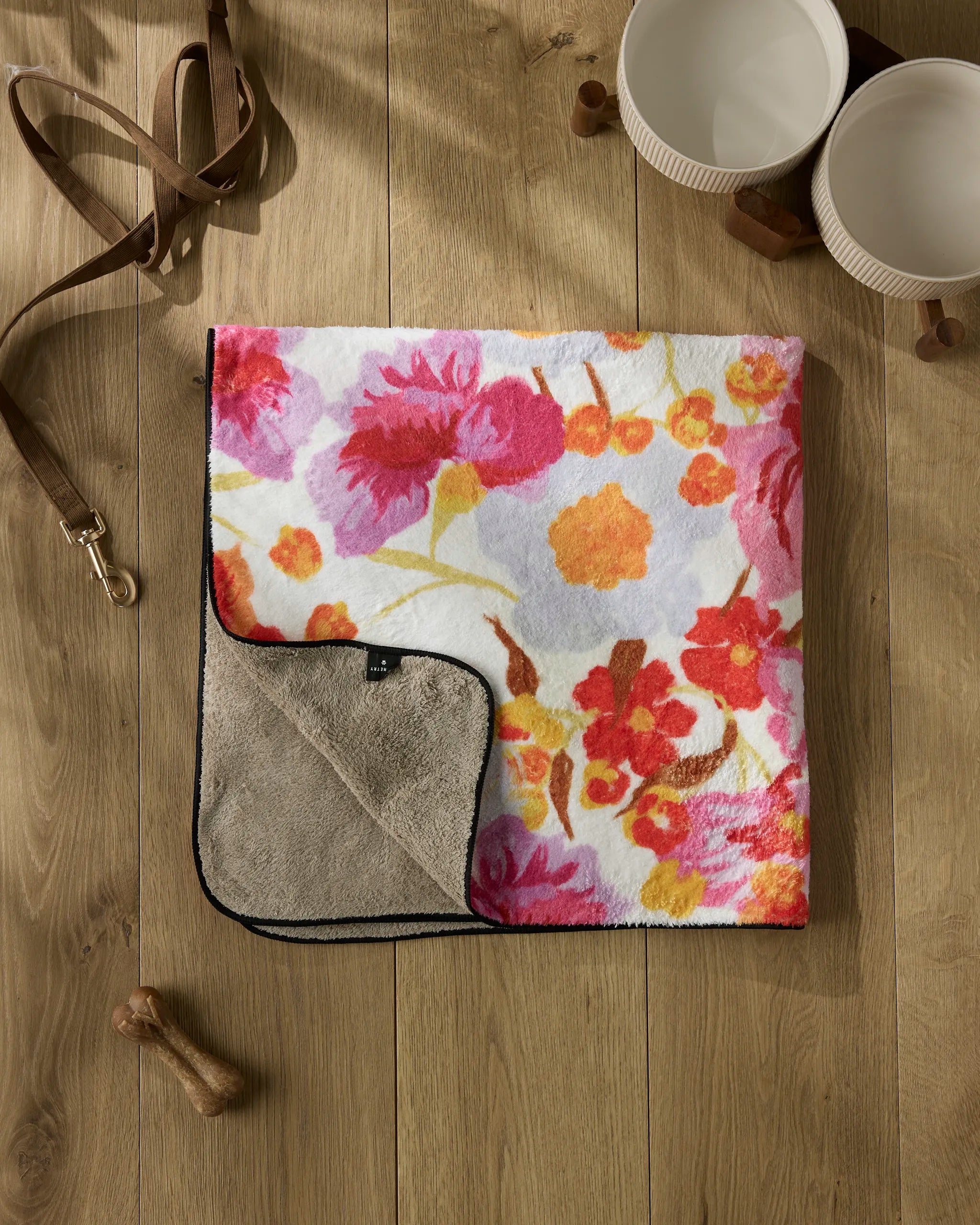 The Geometry Pup Florals dog towel, featuring a pink, orange, and yellow floral pattern, is neatly folded on a wooden floor with two white bowls, a brown leash, and a wooden bone toy nearby.