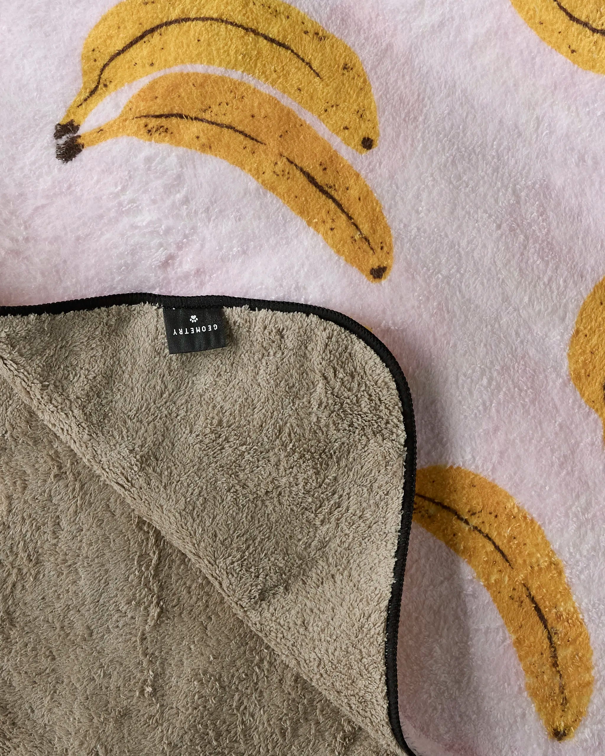The Geometry Pupnana is a soft, plush dog towel blanket with a banana pattern on light pink. A flipped corner reveals its fluffy, light brown underside and a black-bordered CLOVERNY tag—ideal for cuddles or use as an absorbent towel.